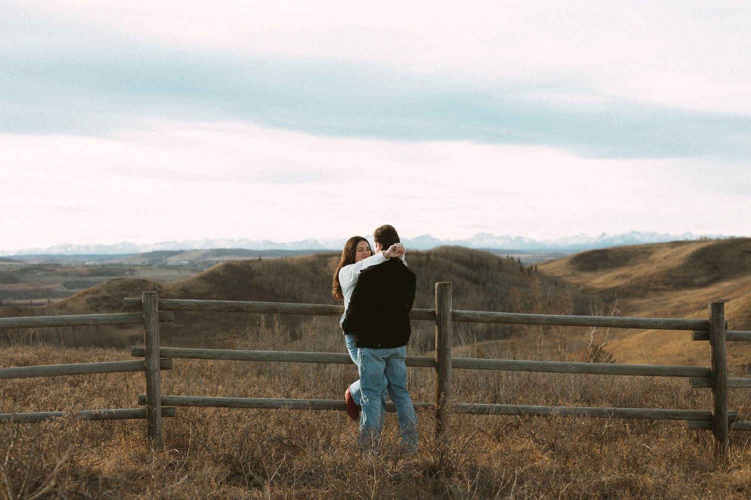 A panoramic view of a rustic wooden fence stretches across brown hills under an overcast sky.