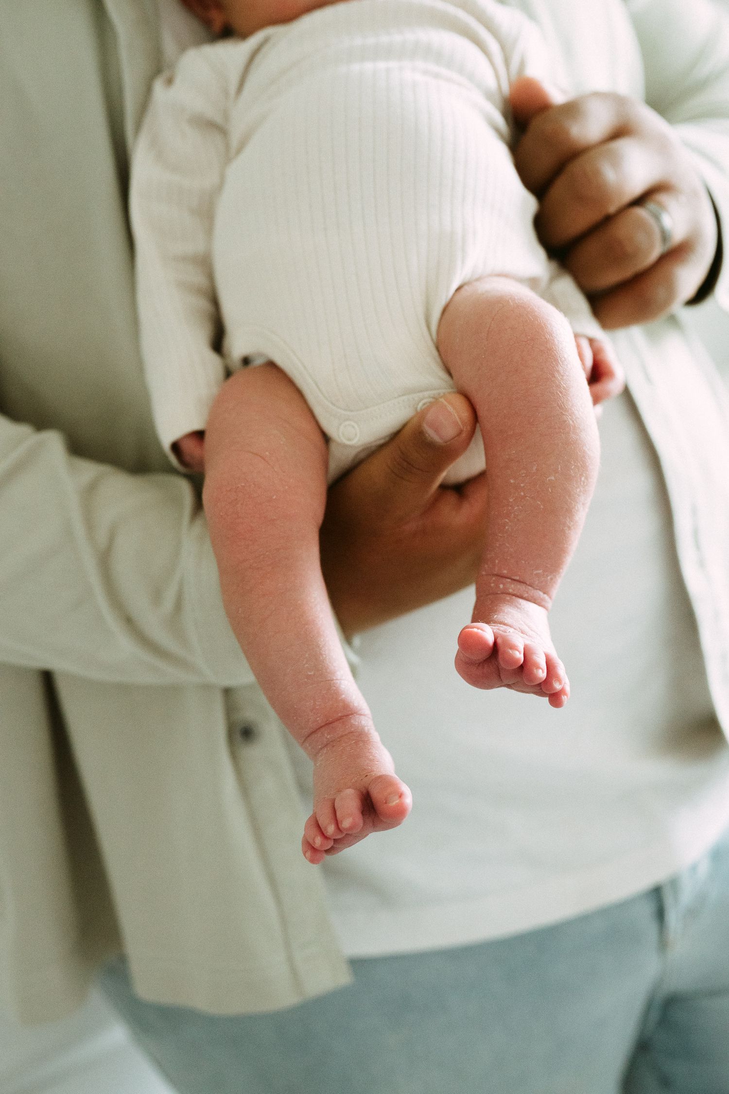 Close-up of a newborn baby being cradled in an adult's arms, wearing a white onesie with tiny feet visible.