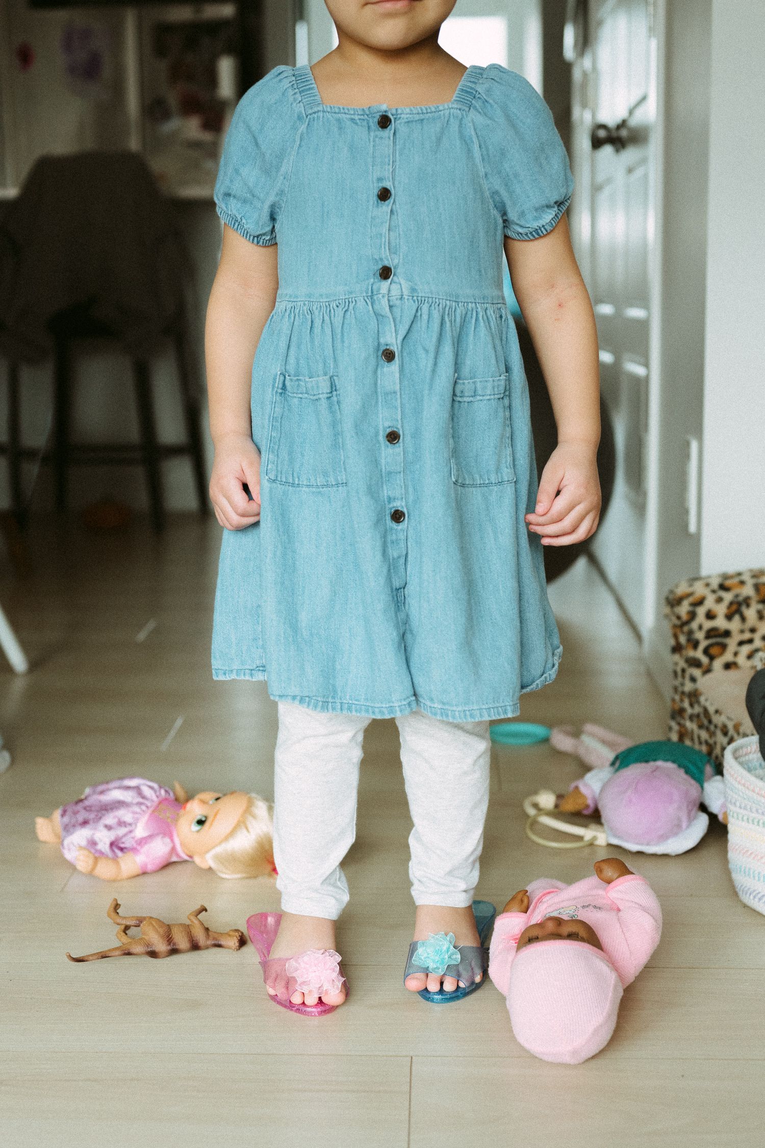 Young girl in denim dress and white leggings stands surrounded by dolls and toys on floor.
