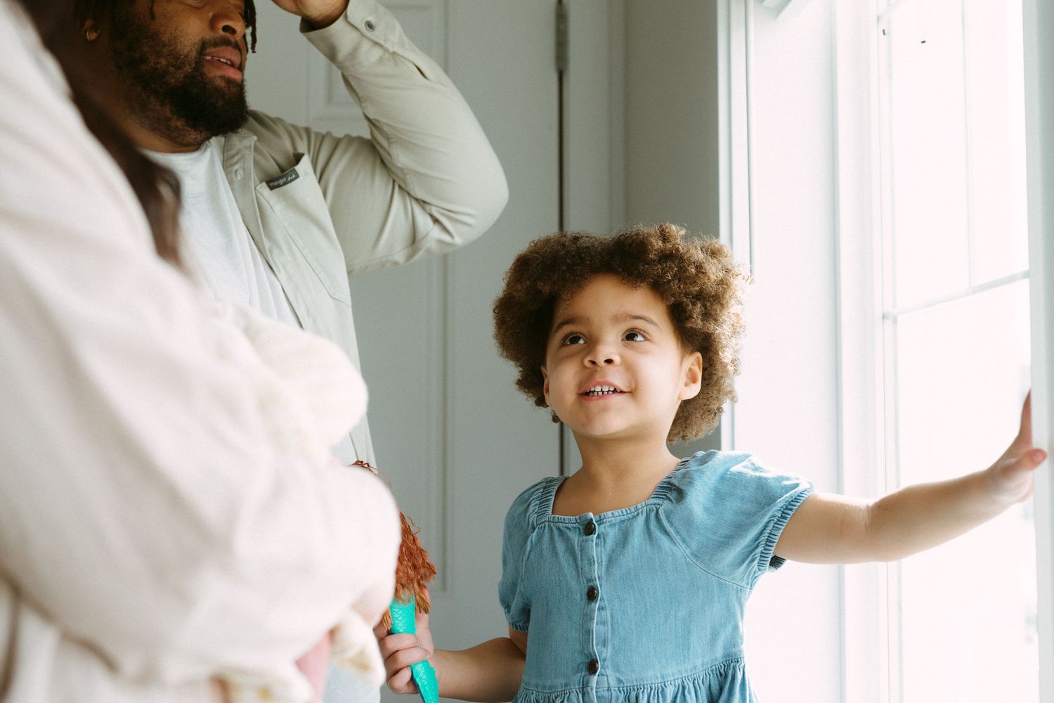 A young child in a blue dress looks up curiously near a window while adults stand nearby indoors.