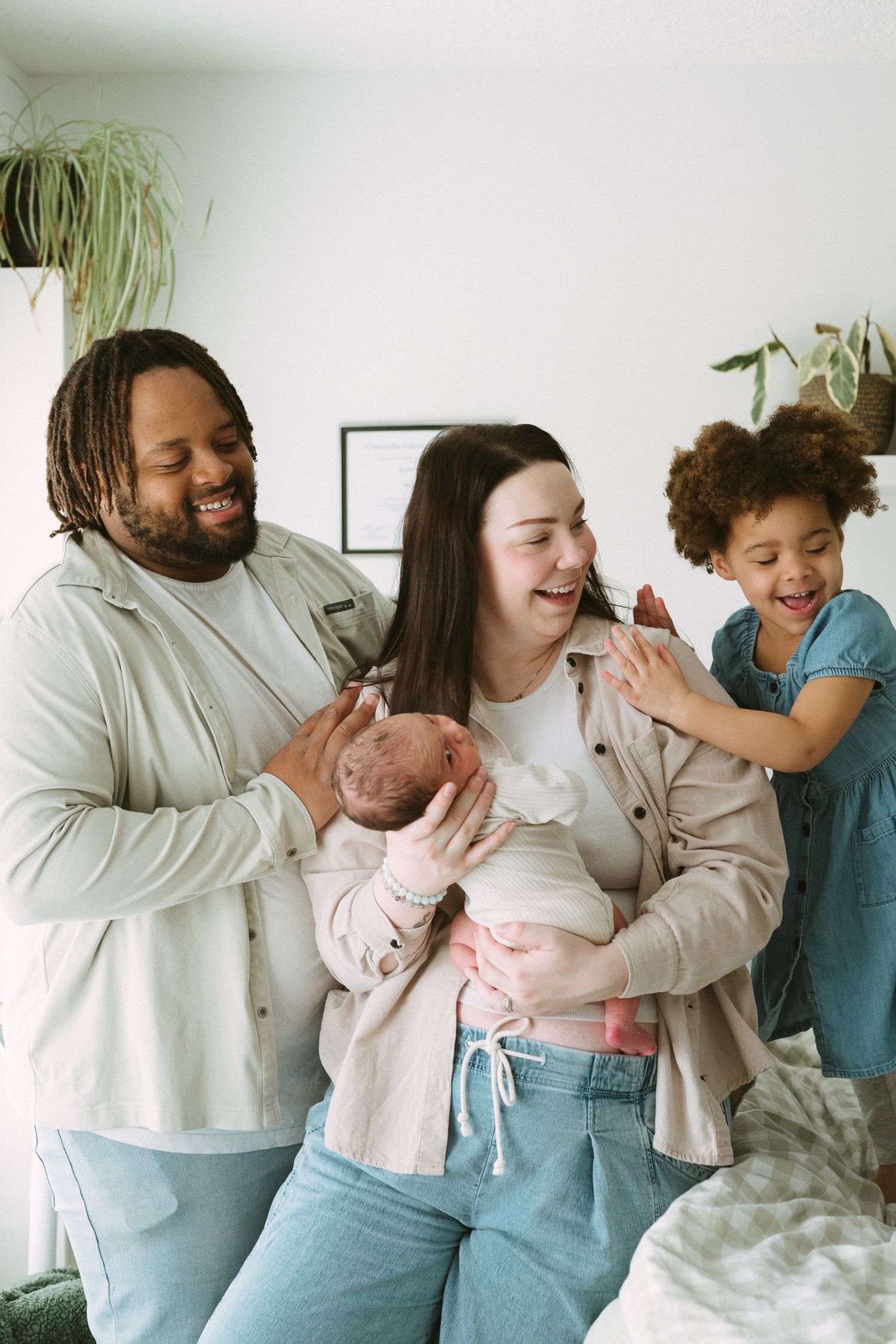A joyful multiracial family laughs together as the mother holds their newborn baby with dad and daughter nearby.
