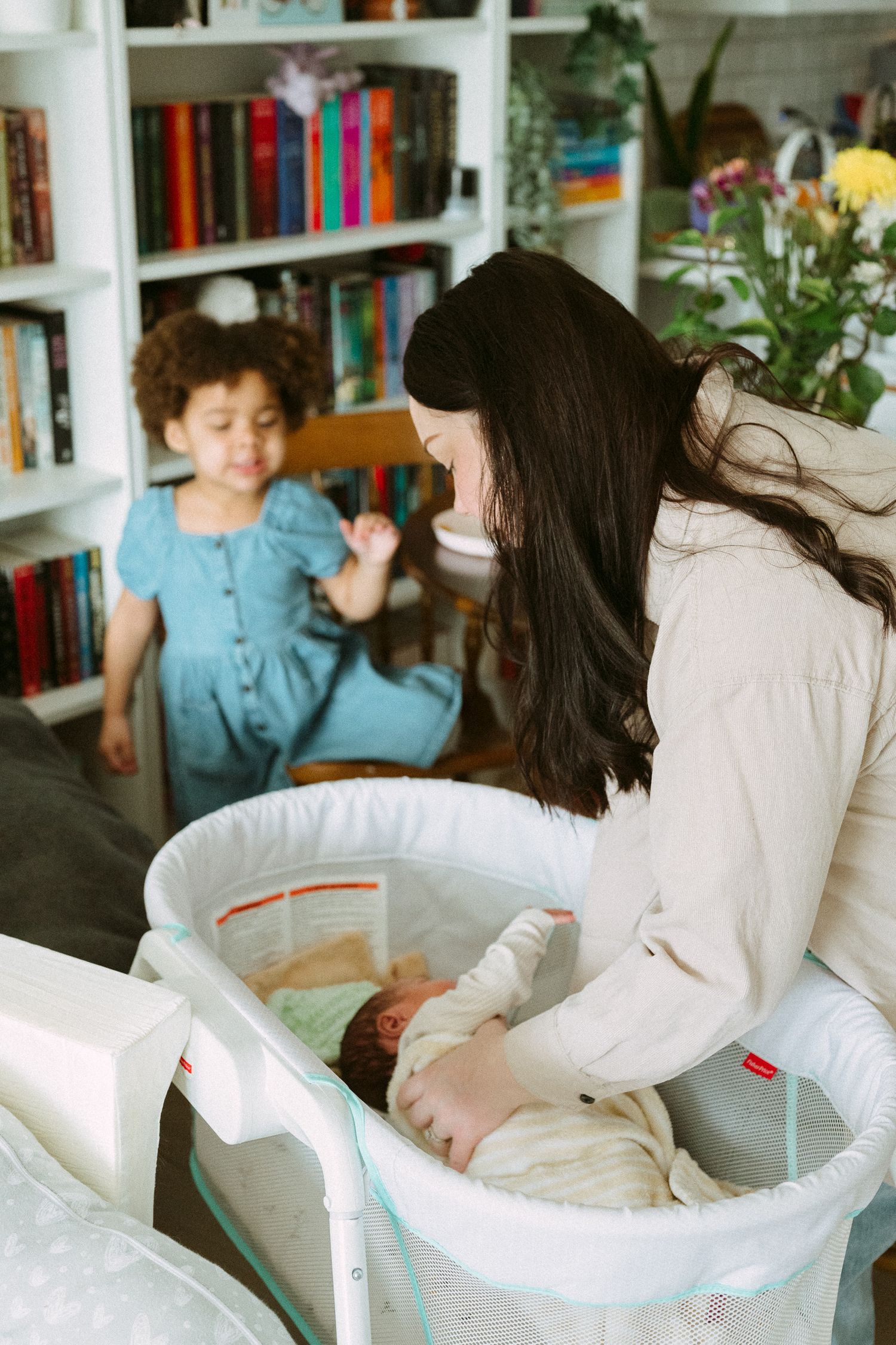 A dark-haired woman tends to a newborn in a white bassinet as a young curly-haired girl looks on.
