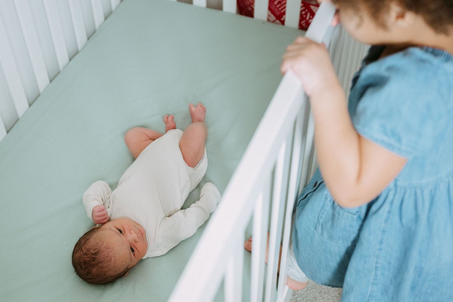 A toddler girl in a blue dress stands beside a white crib with a newborn baby inside.