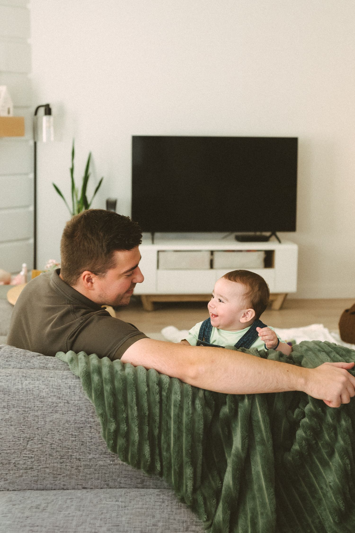 Two people relaxing together on a gray couch with a green blanket in a bright living room.