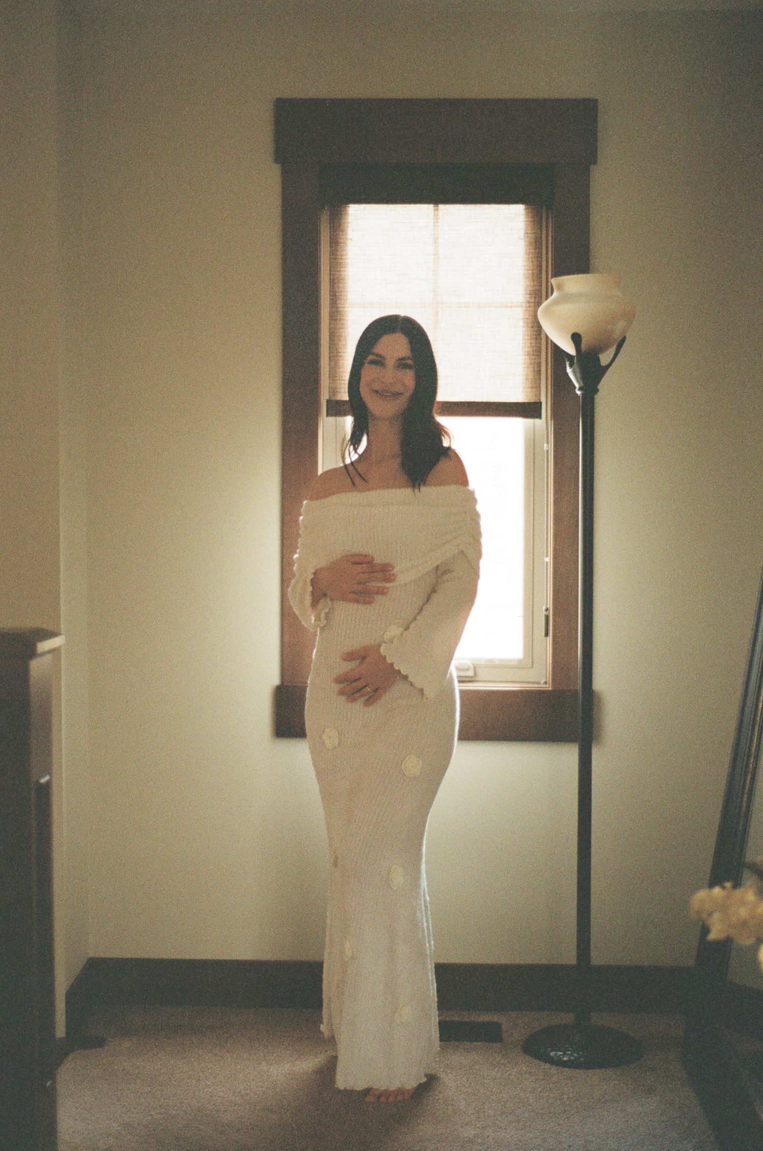 Pregnant woman in white off-shoulder maxi dress standing by sunlit window, cradling baby bump indoors.