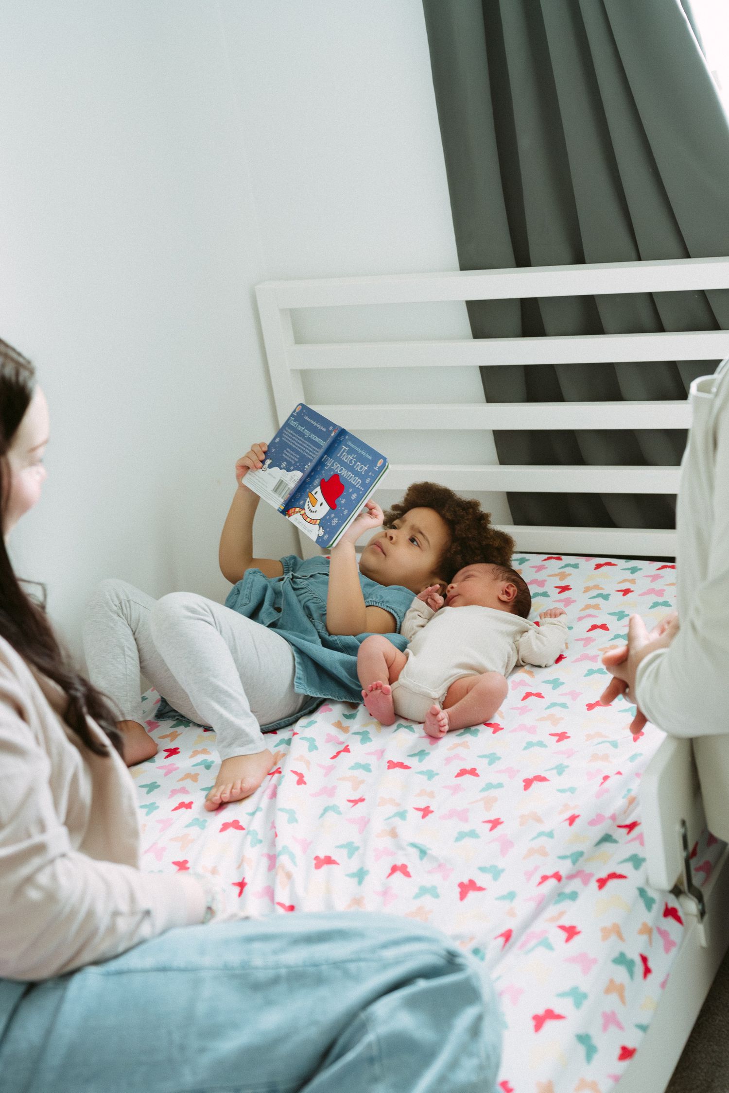 Young toddler reading a picture book on a floral bedsheet beside a newborn baby while mother watches nearby.