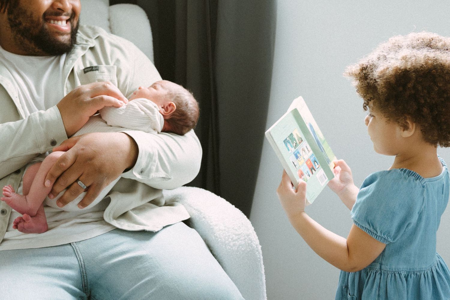 Father holding newborn baby while young toddler daughter shows him a picture book.