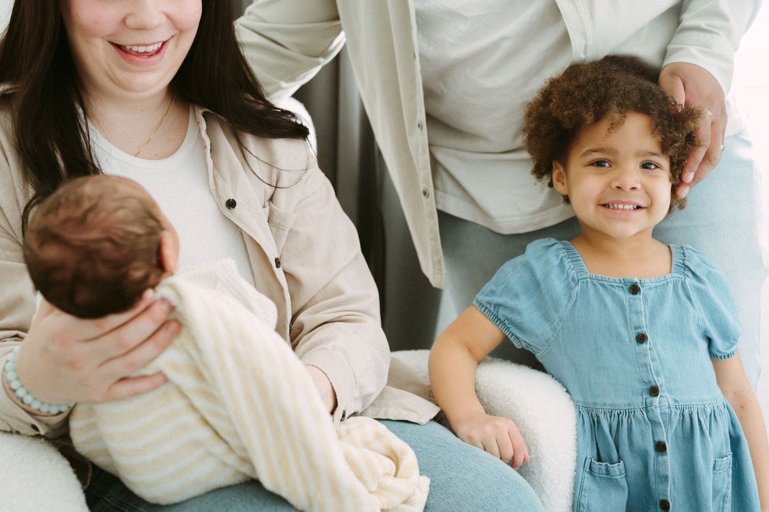 A mother holds her newborn baby while her curly-haired toddler daughter smiles beside her.