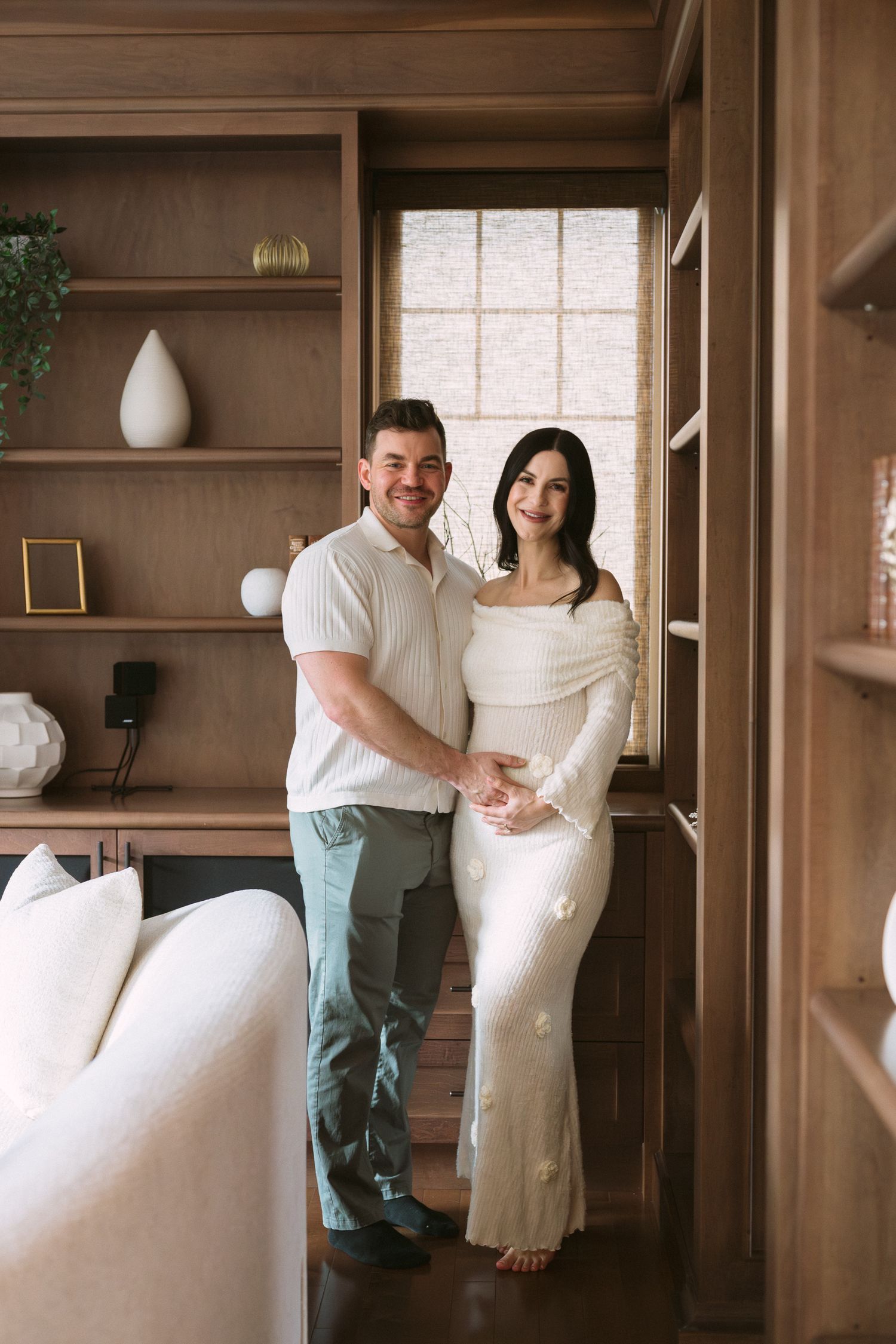 Smiling couple posing together in wood-paneled room, pregnant woman in white dress with partner in casual attire.