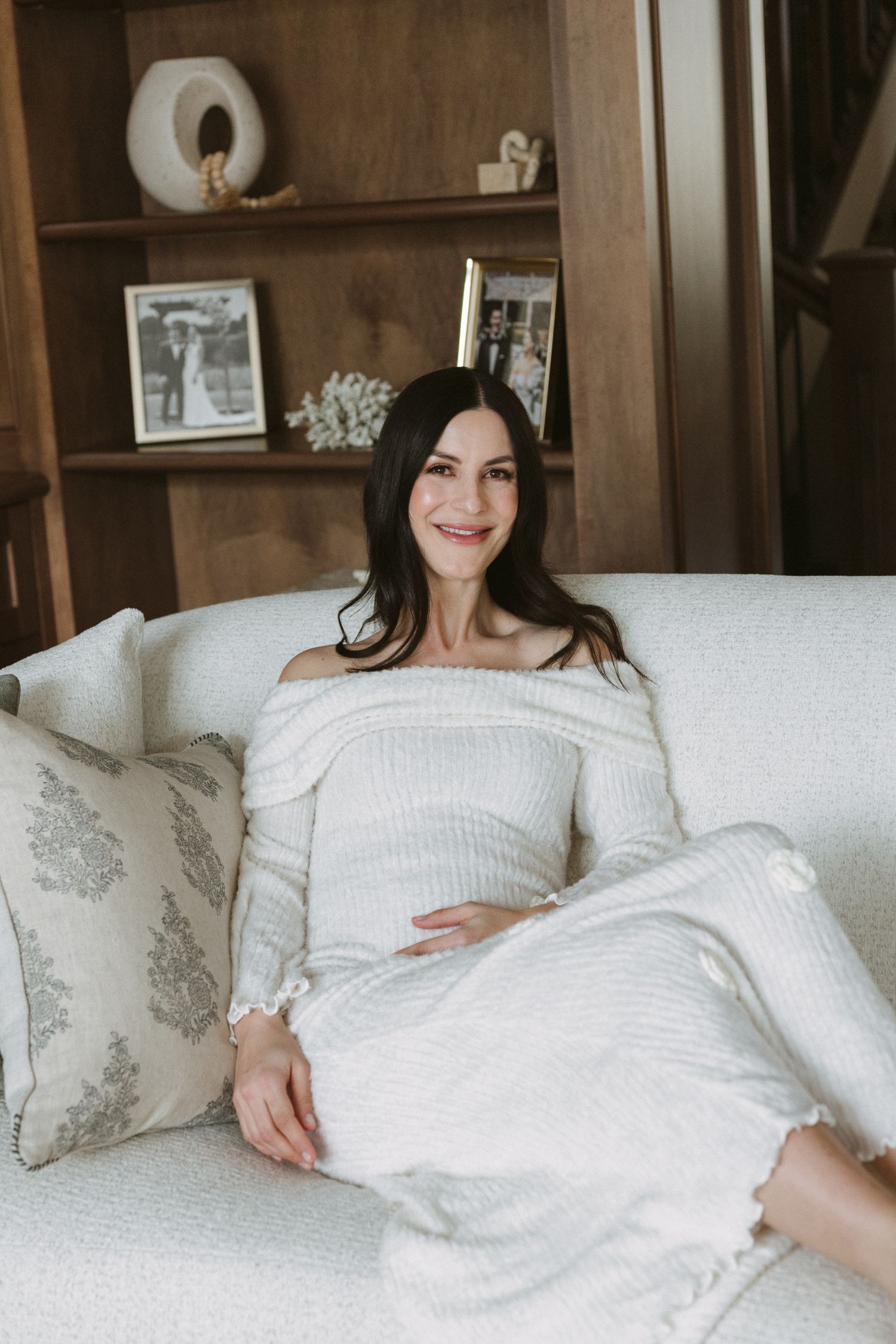 Smiling brunette woman in white off-shoulder dress sitting on cream sofa with decorative pillows in cozy living room.