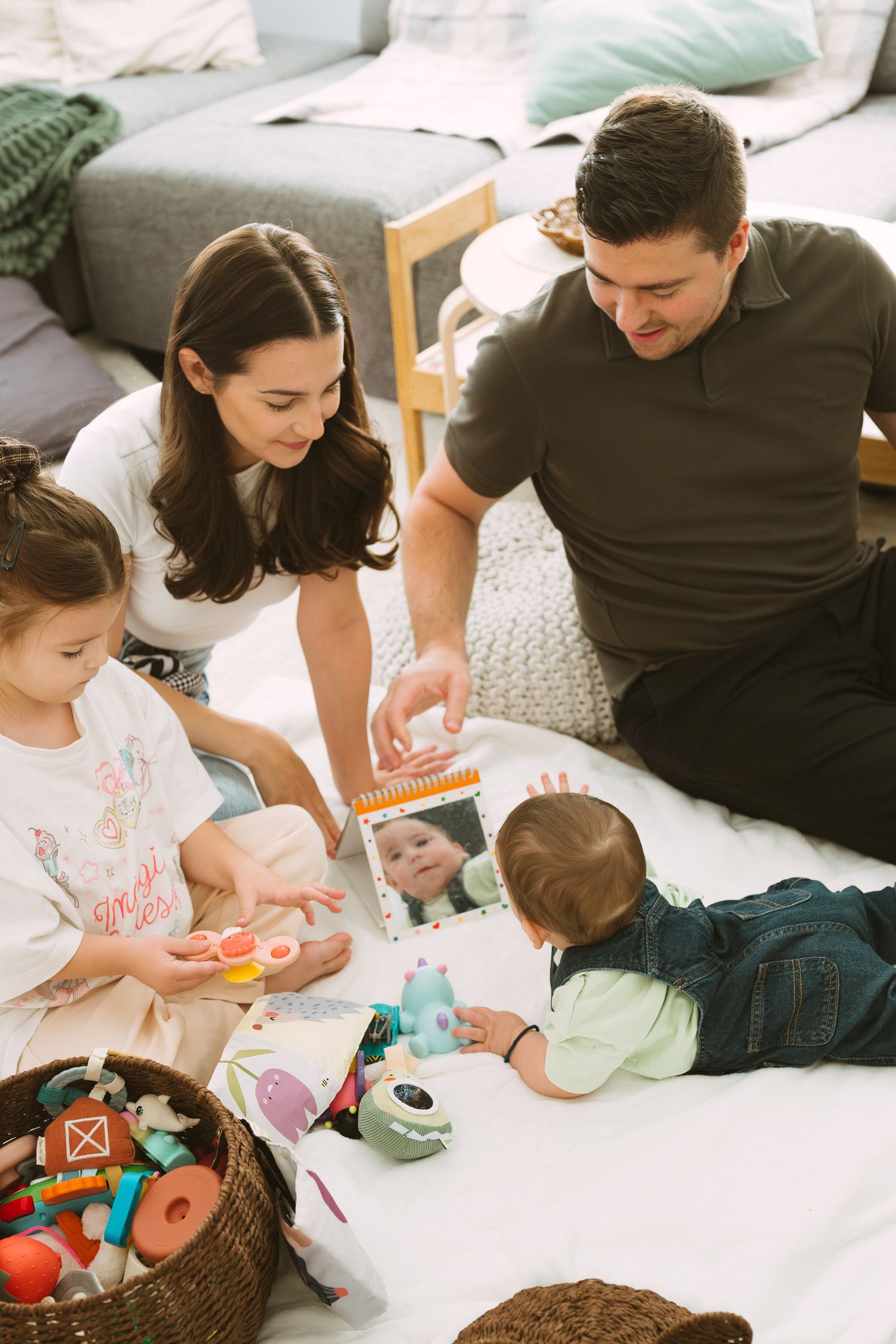 A family sits together on a white bed reading books and playing with colorful toys from a basket.