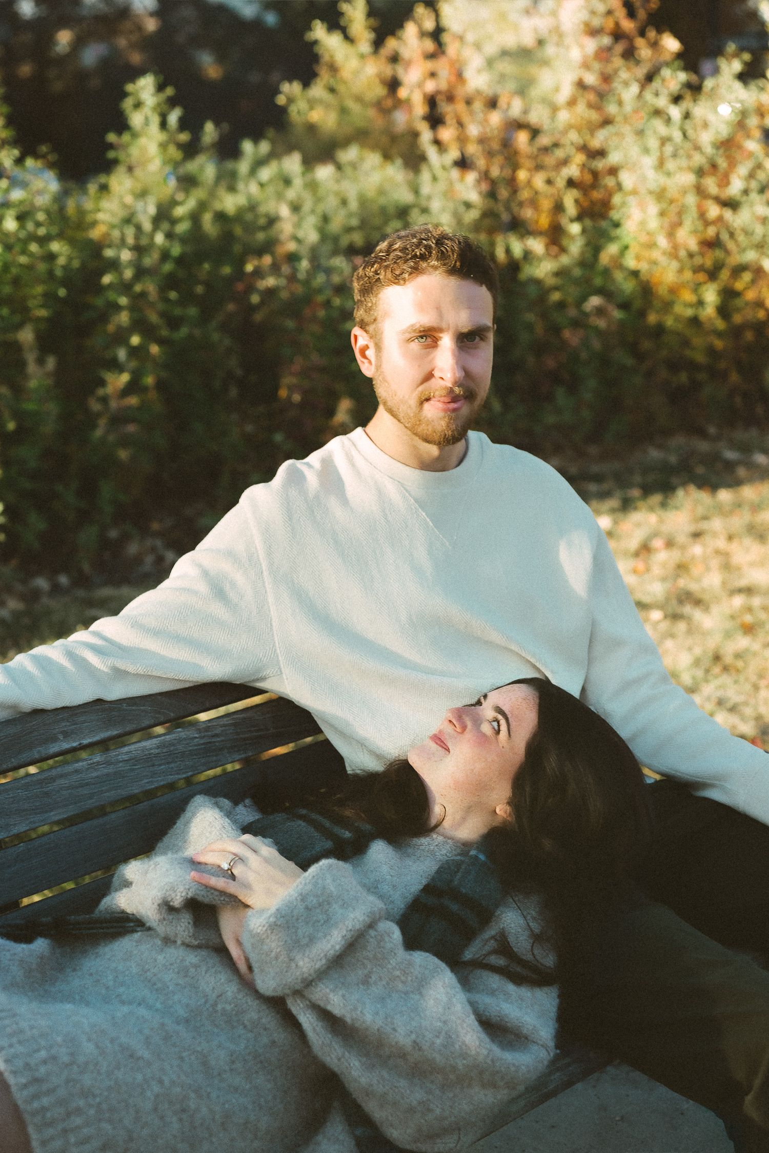 Two people share a tender moment while resting together on a wooden bench in golden sunlight.