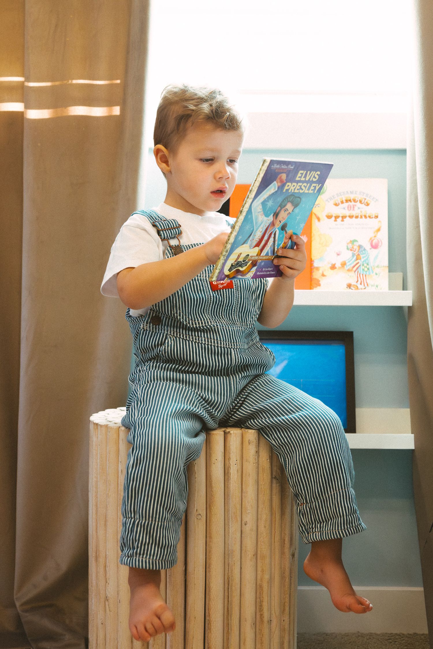 A young child in striped overalls sits barefoot on a wooden surface while reading a book near a bookshelf.