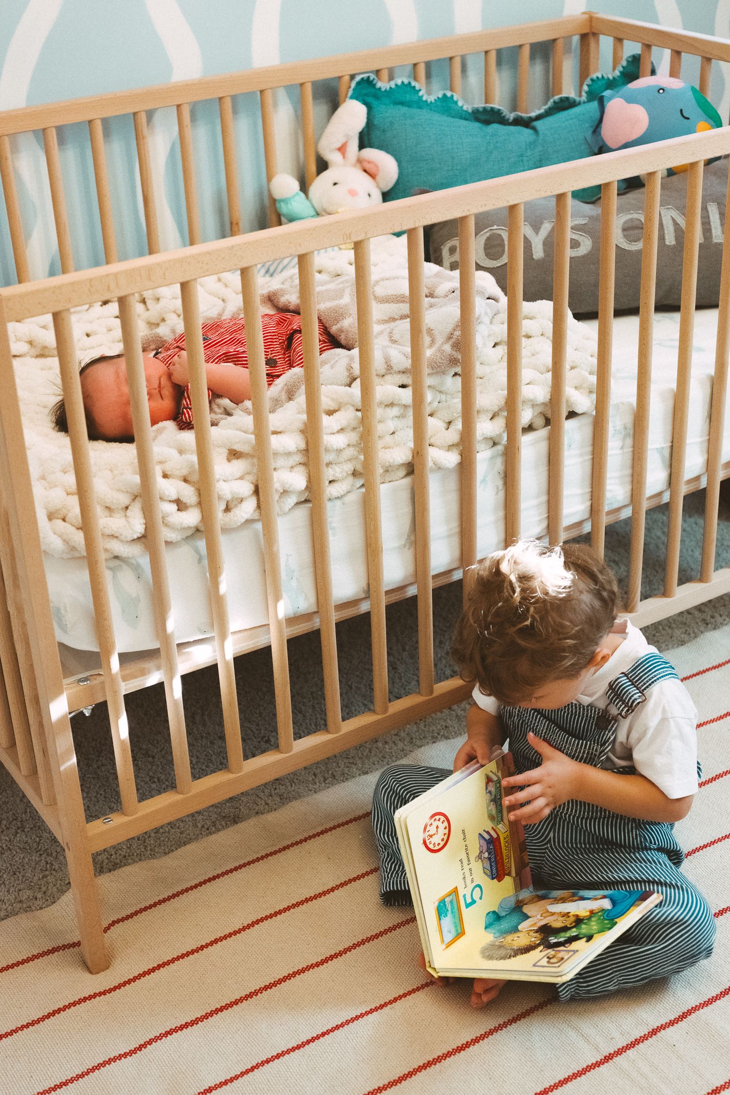 Young child sitting on floor reading a picture book against wooden railing in a casual indoor setting.