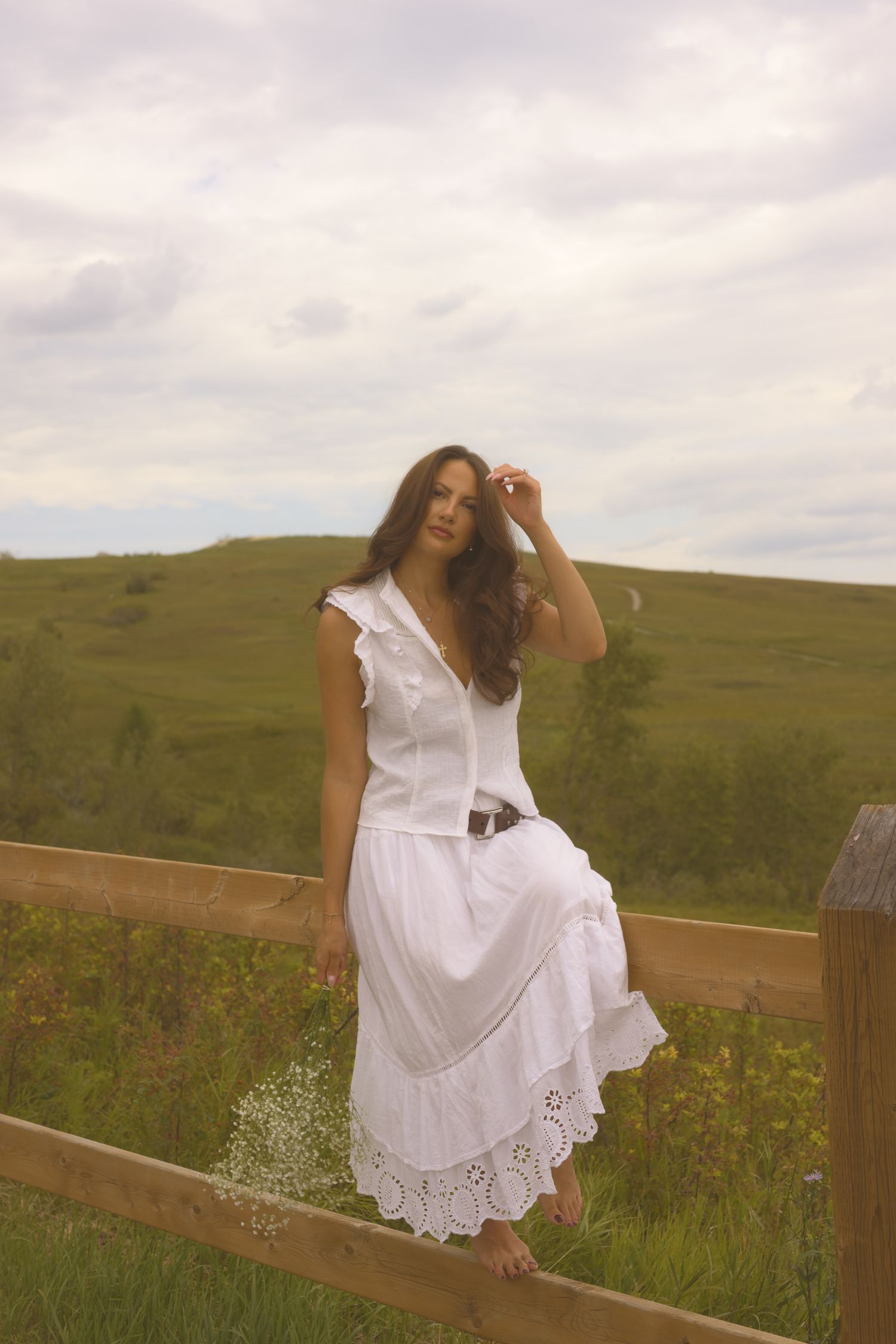 A dreamy portrait on a wooden fence showcases a flowing white eyelet dress against rolling hills.
