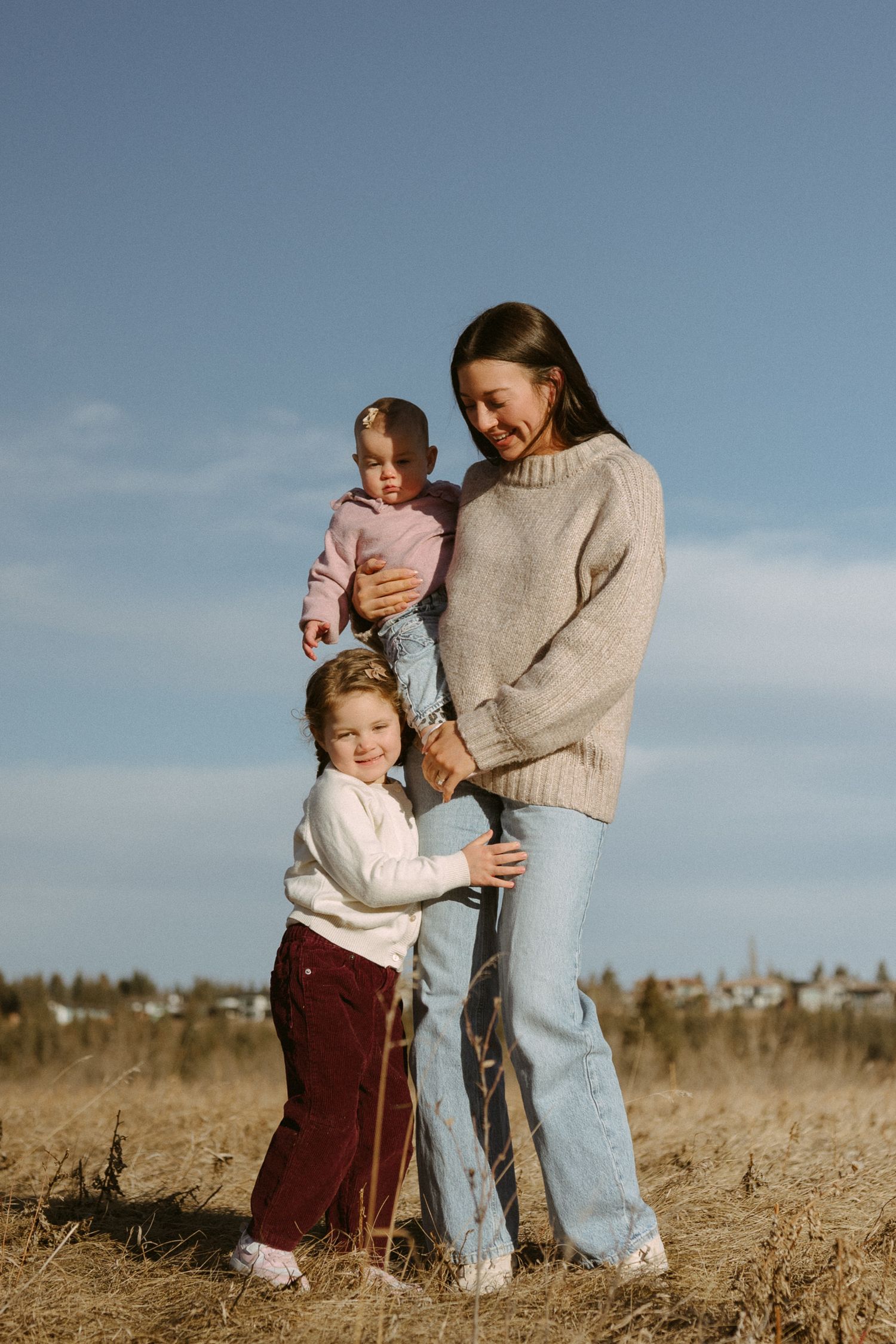A family in casual sweaters and jeans stands together in a sunny field during golden hour.
