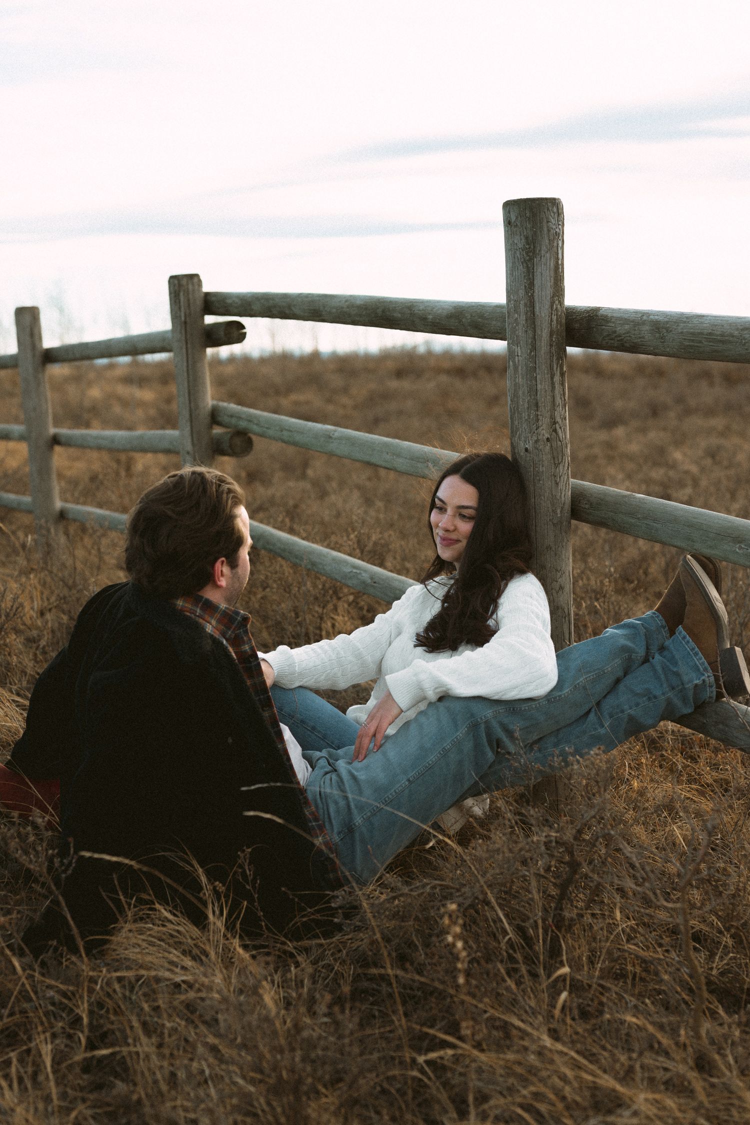 Couple relaxing together against a wooden fence in a golden field during sunset.