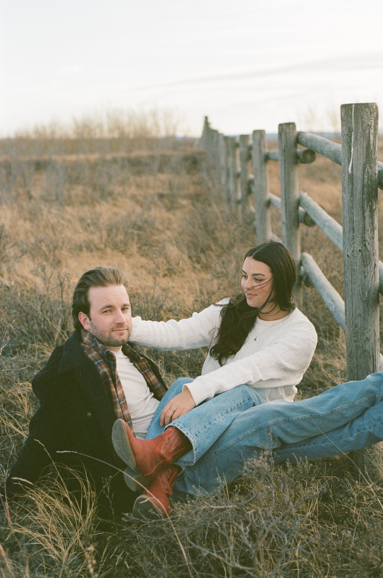 A couple relaxes together on the grass near a wooden fence in a rustic outdoor setting.
