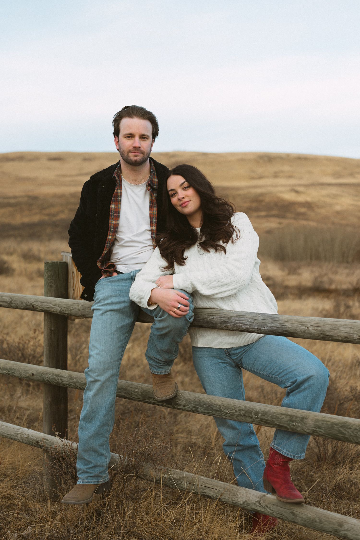 Couple in casual attire sharing an intimate moment while sitting on a wooden fence in a rural landscape.