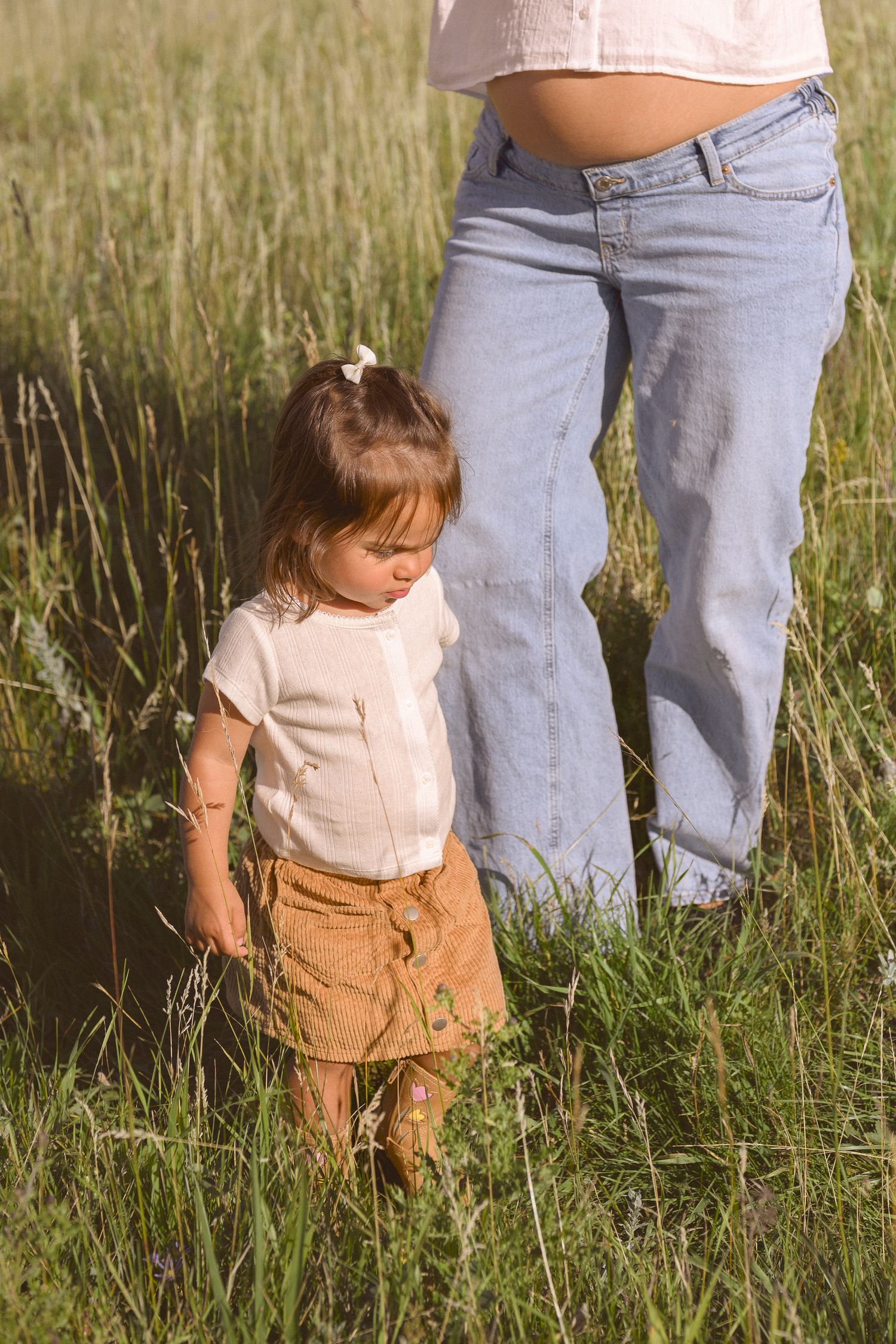 A small child in a white top and brown skirt plays in a grassy meadow on a sunny day.