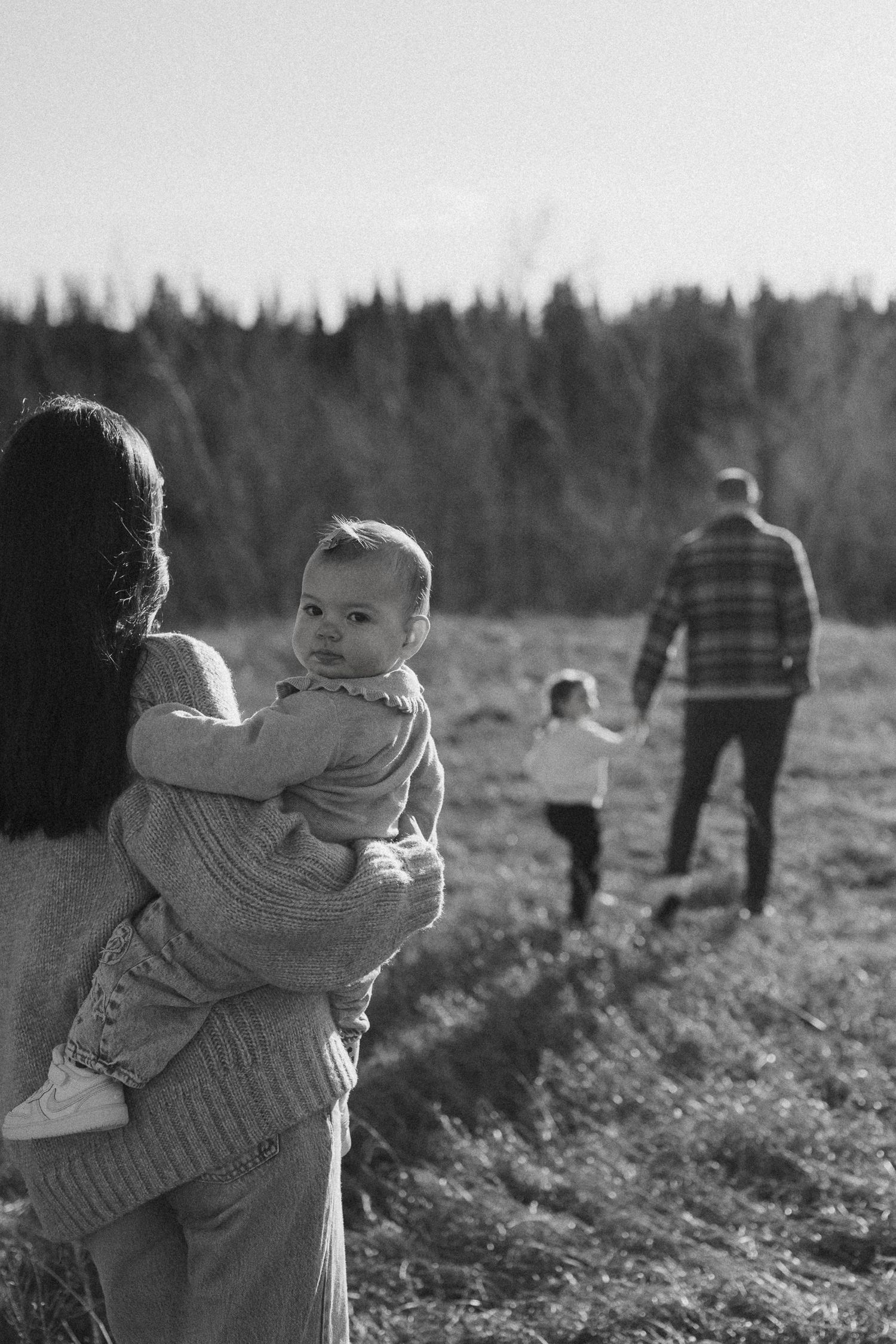 A tender black and white scene of a child being carried in a field with evergreen trees in the background.