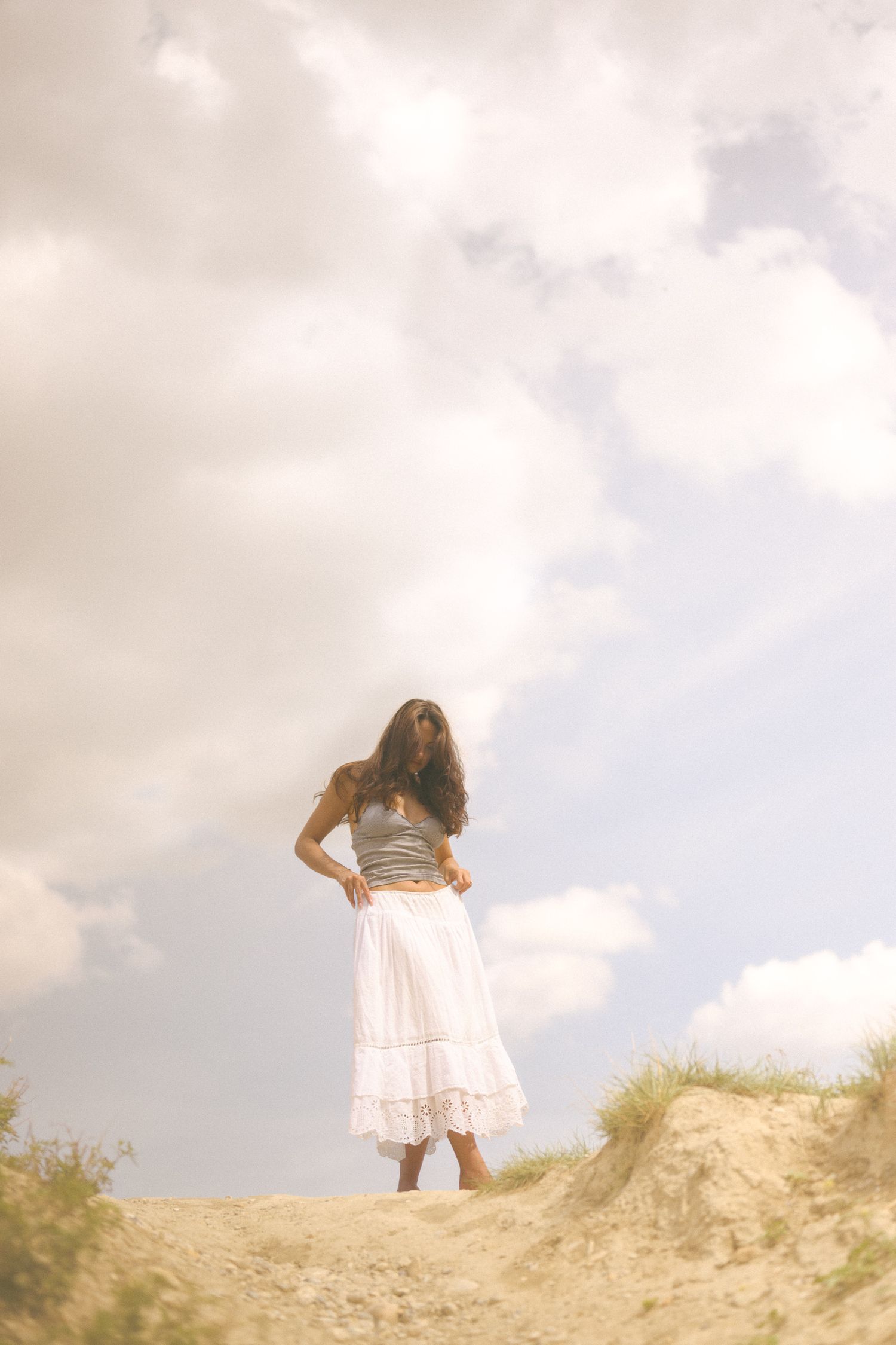 Someone stands atop a sandy hill wearing a white dress and gray top.