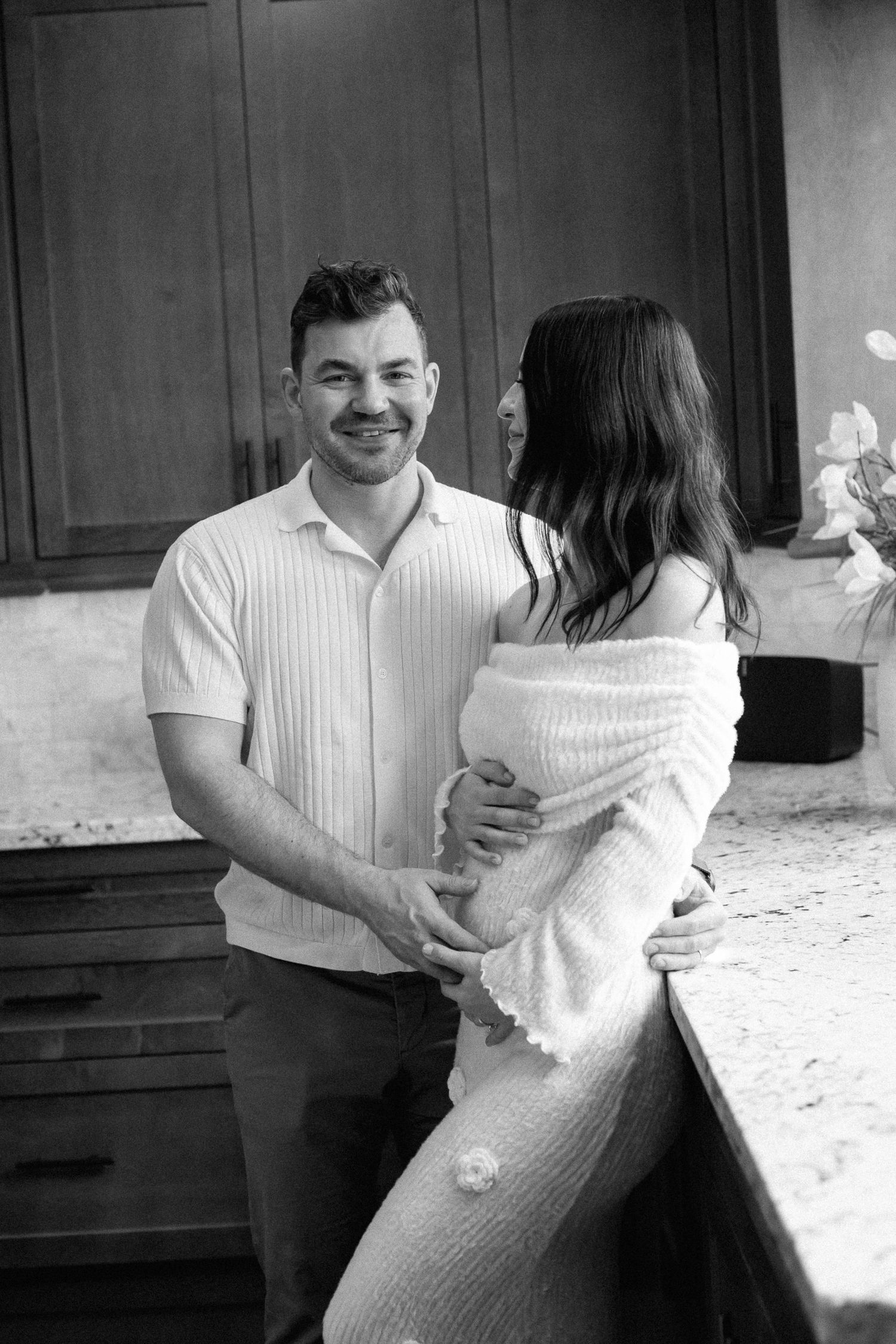 Black and white photo of a smiling man holding a pregnant woman's belly in a kitchen setting.