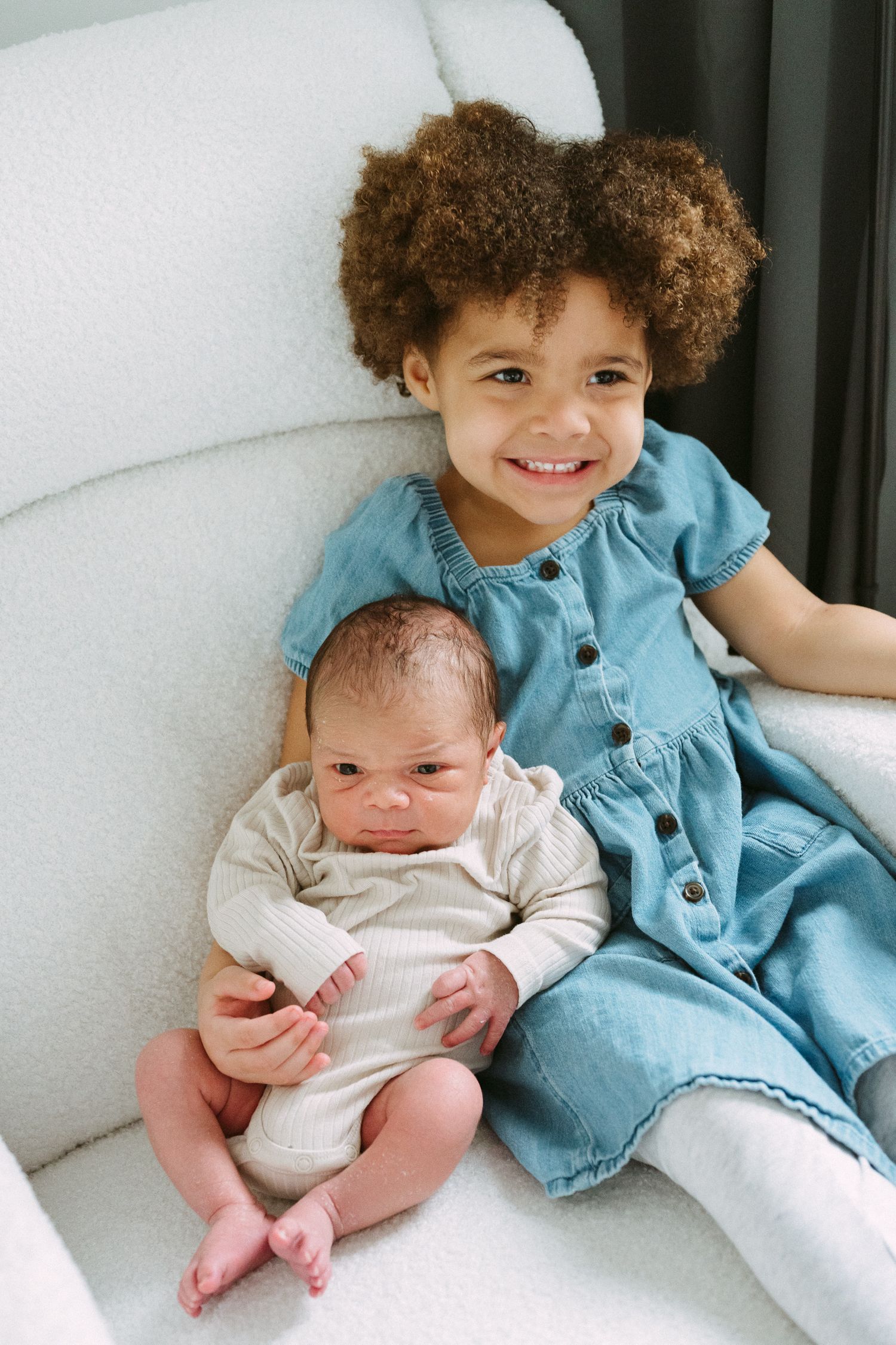 Young girl in denim dress smiling while holding newborn baby sibling on white chair.