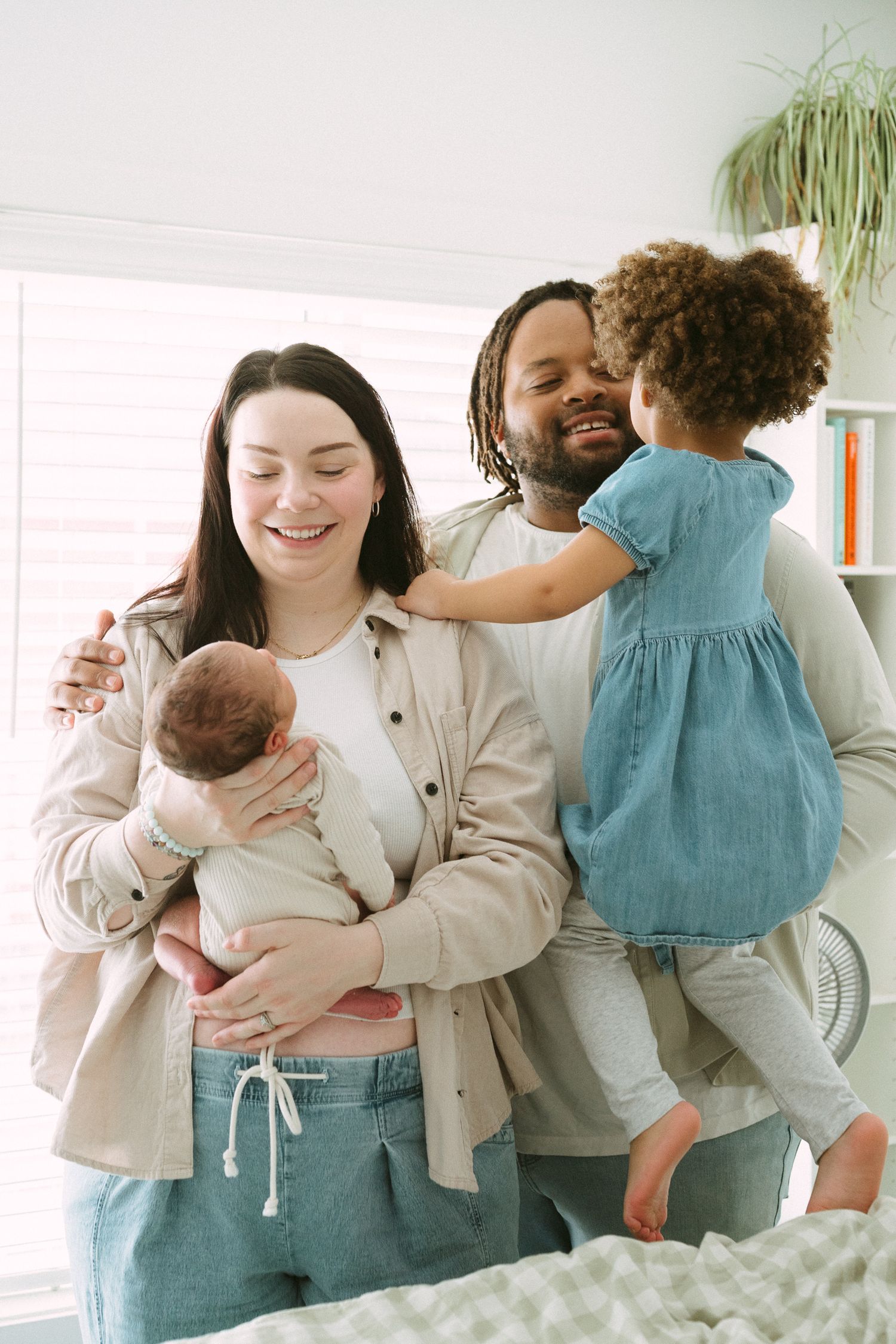 Happy interracial family with parents holding newborn baby and toddler daughter in bright bedroom.
