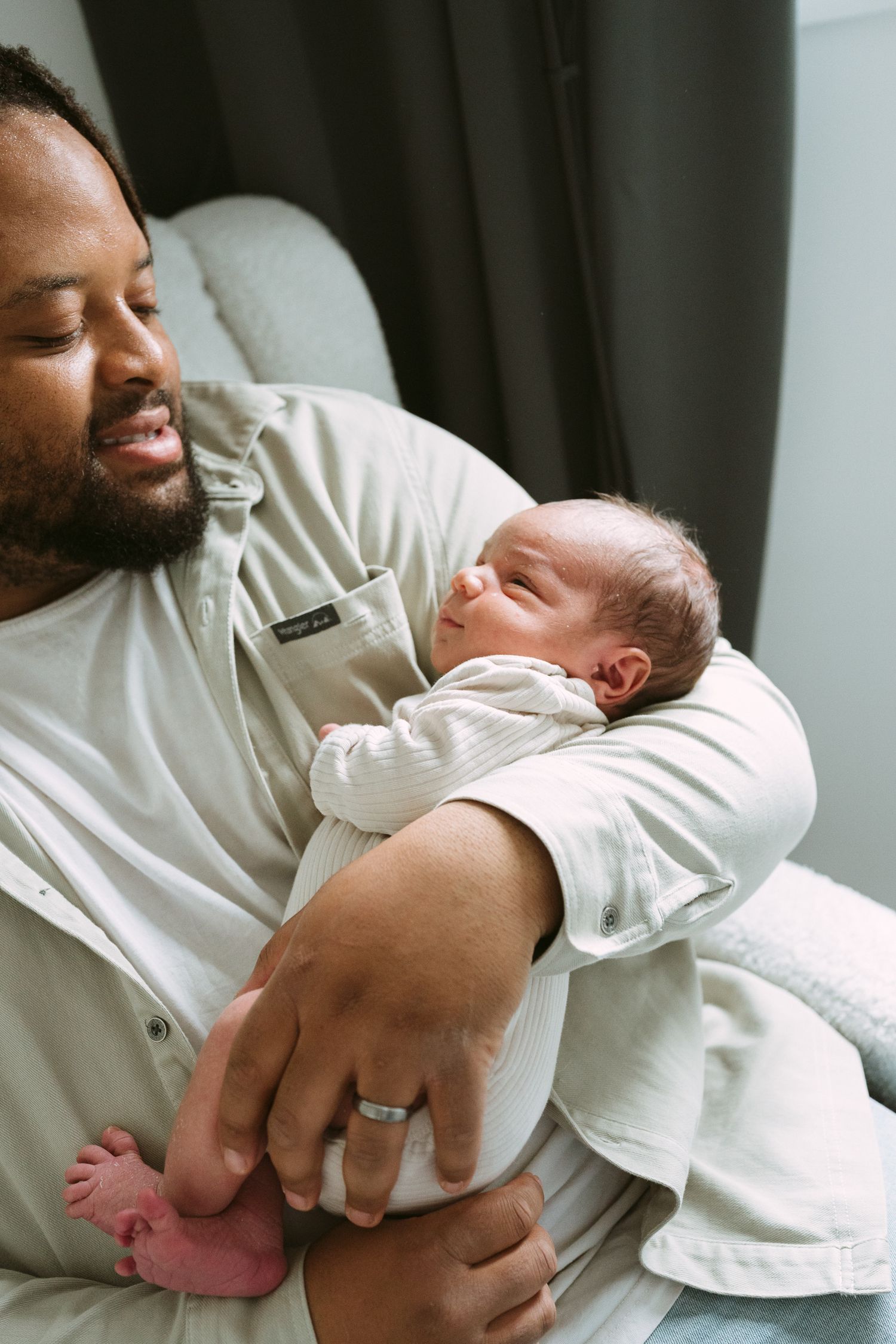 A Black father sits in a chair cradling his sleeping newborn baby in his arms.