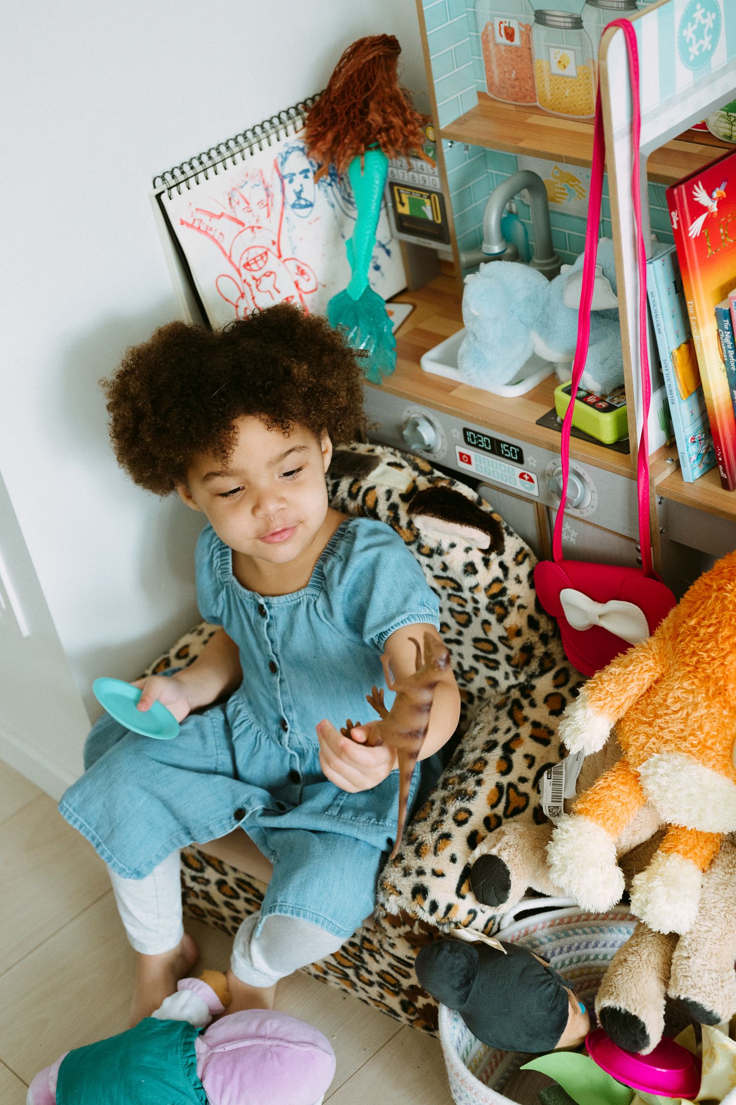 Young girl in denim jumpsuit playing with toys surrounded by stuffed animals and books in a colorful playroom.