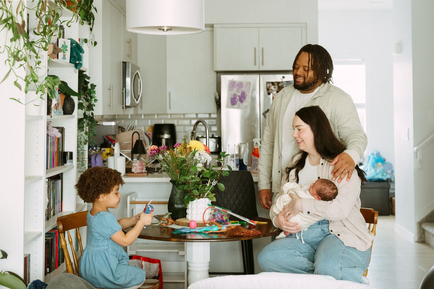 A happy family gathers in a bright kitchen, parents with a newborn and toddler sharing a warm moment together.