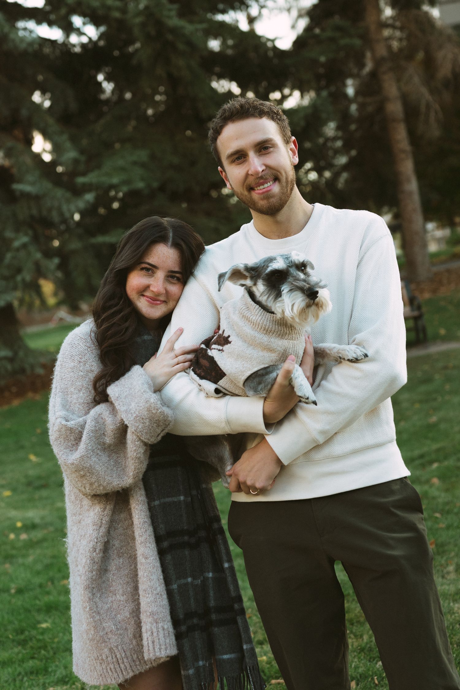 A couple poses with their small white dog in a park setting during a casual outdoor photoshoot.
