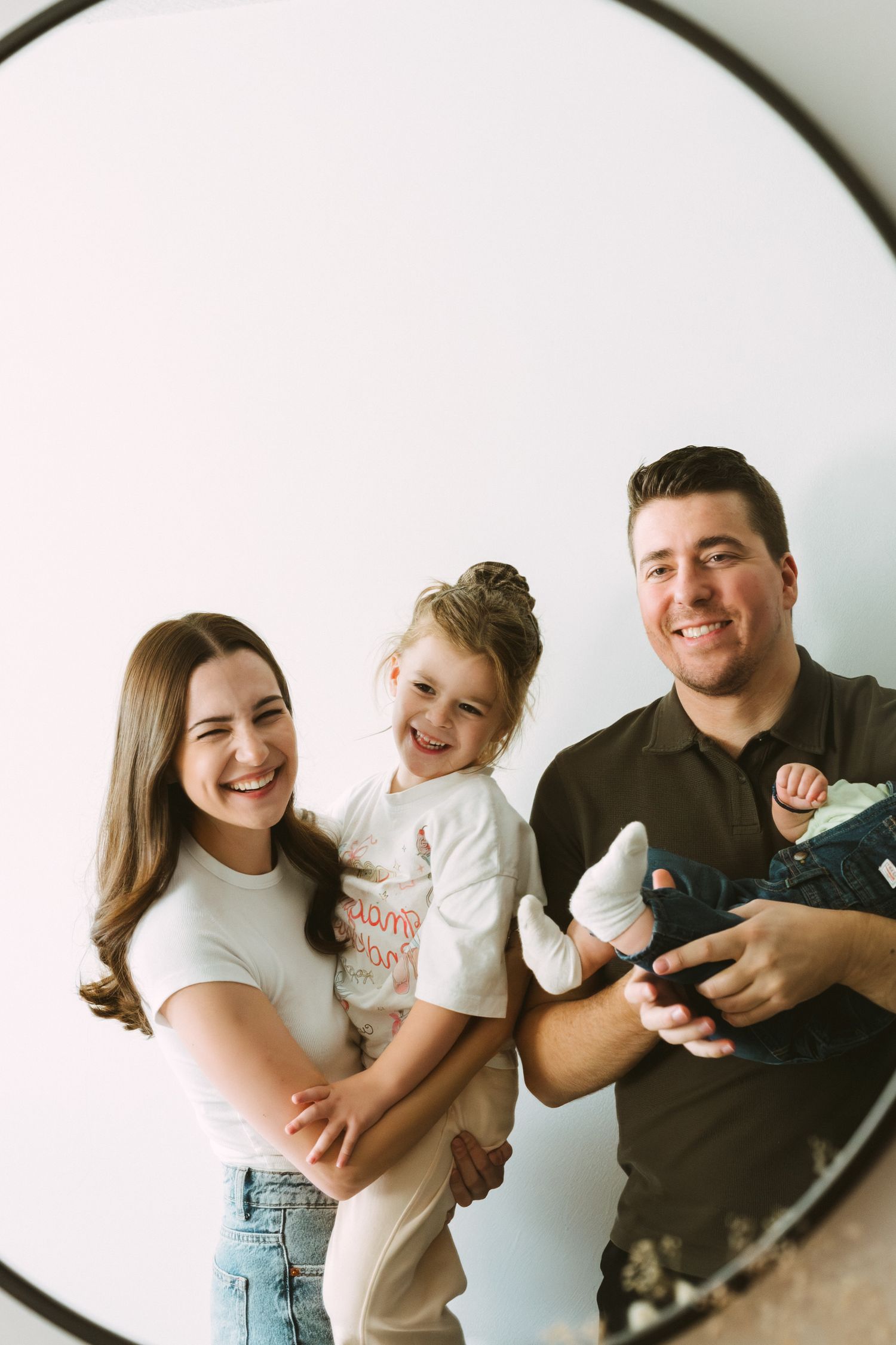 A happy family laughing and playing together against a white wall in matching casual outfits.