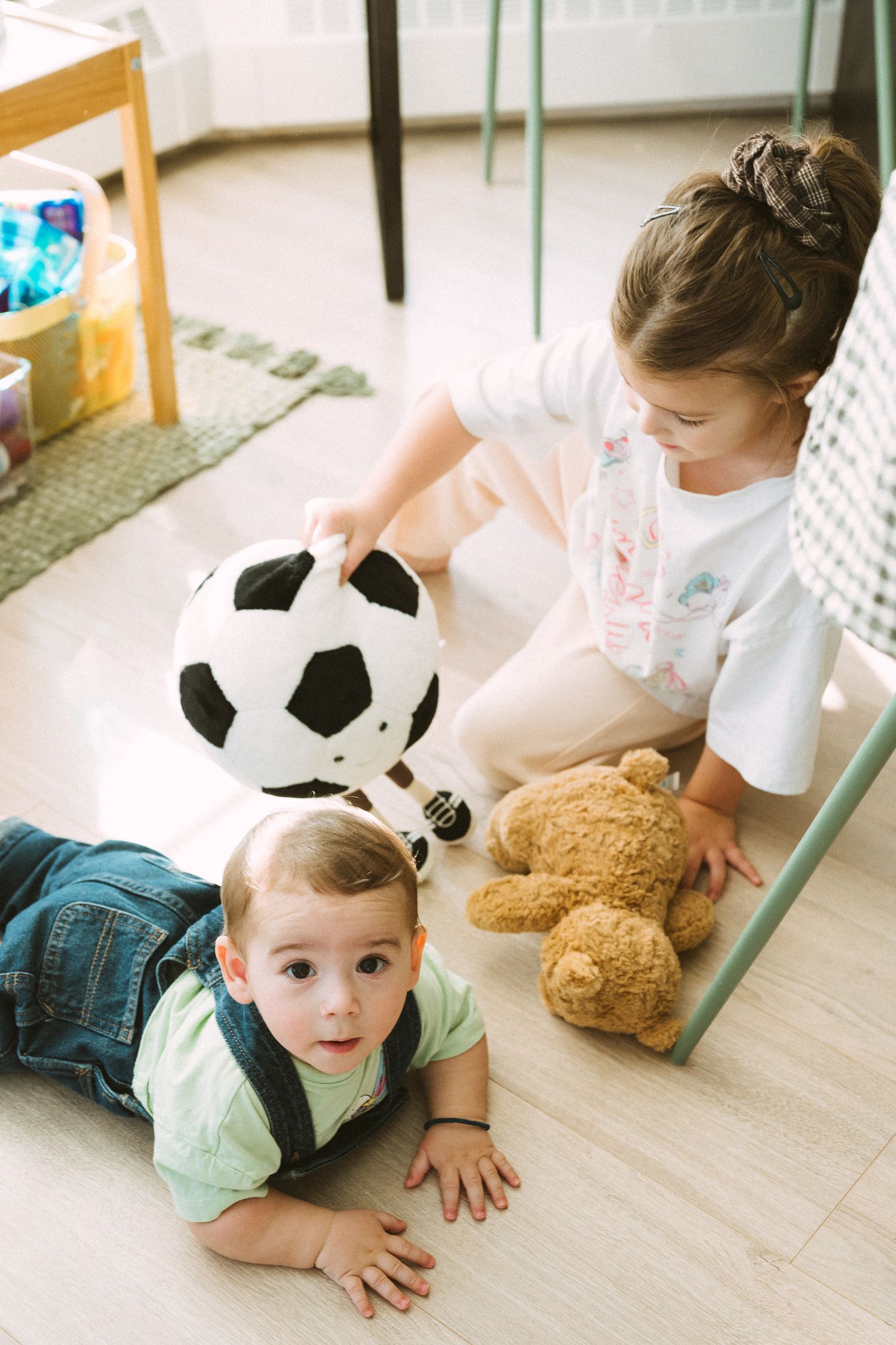 Children playing on the floor with a soccer ball and teddy bear in a bright indoor setting.