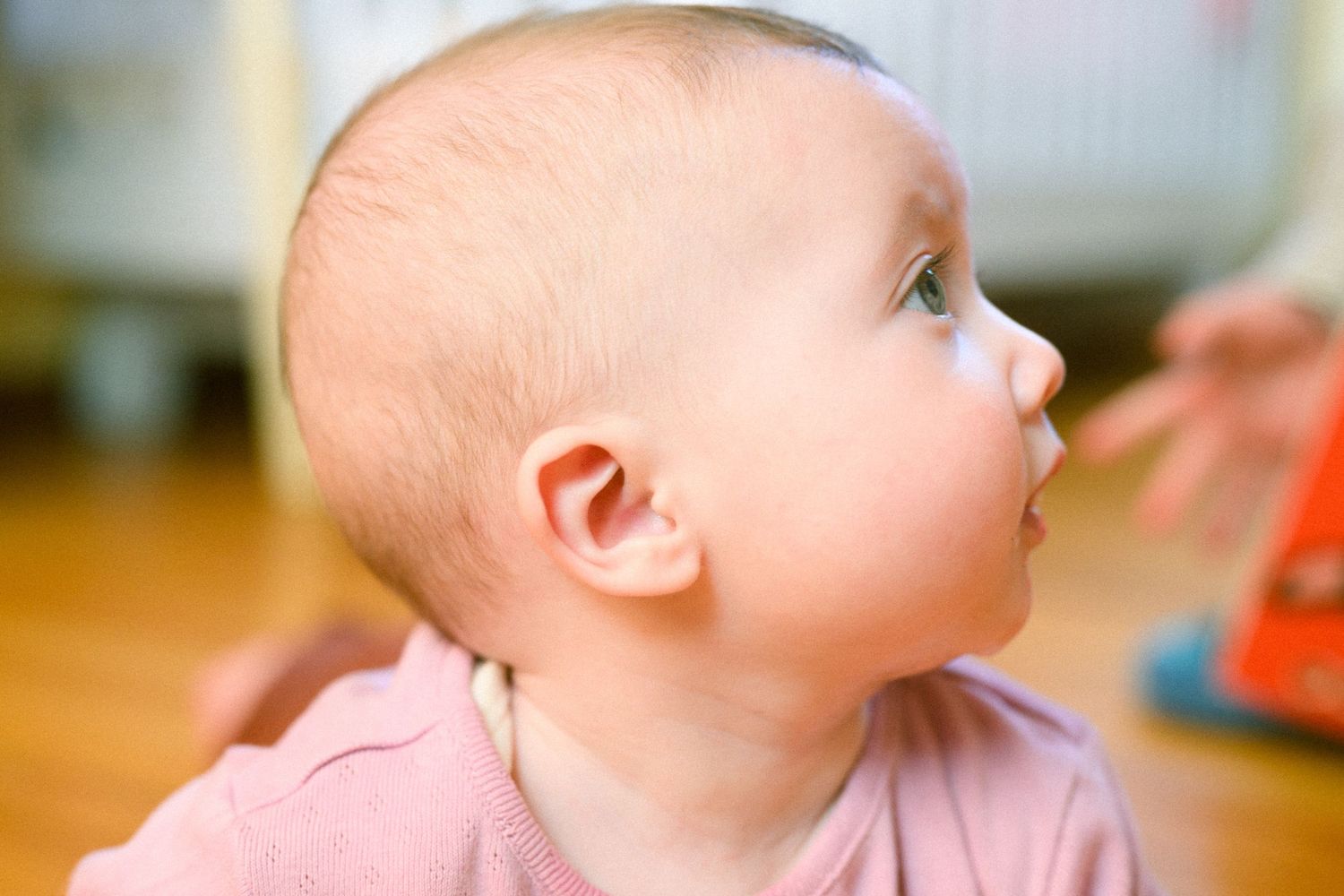 Baby in pink clothing gazes upward with a curious expression on a wooden floor.