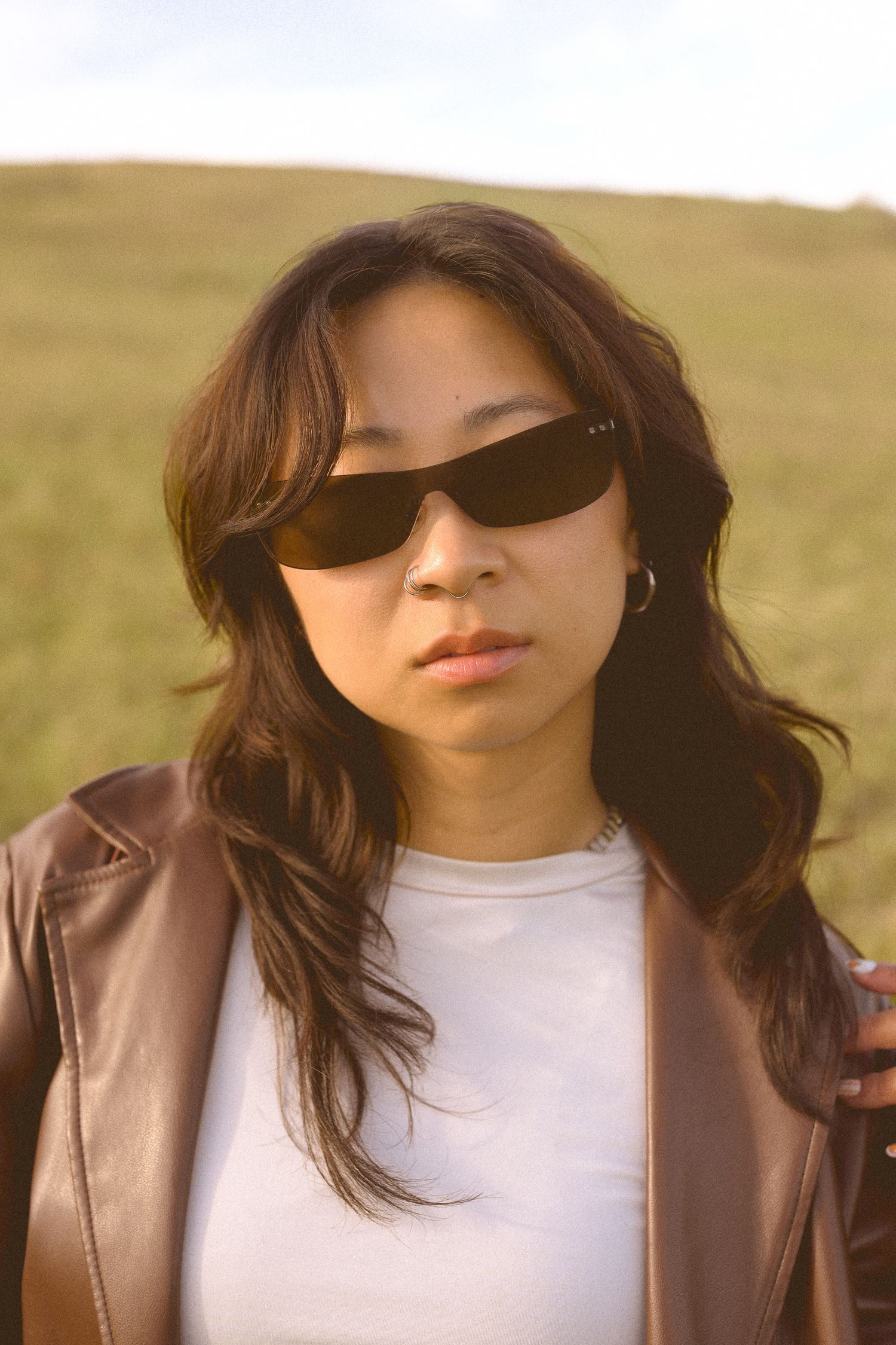 A stylish pose in sunglasses, white top and brown leather jacket against a golden hour backdrop.