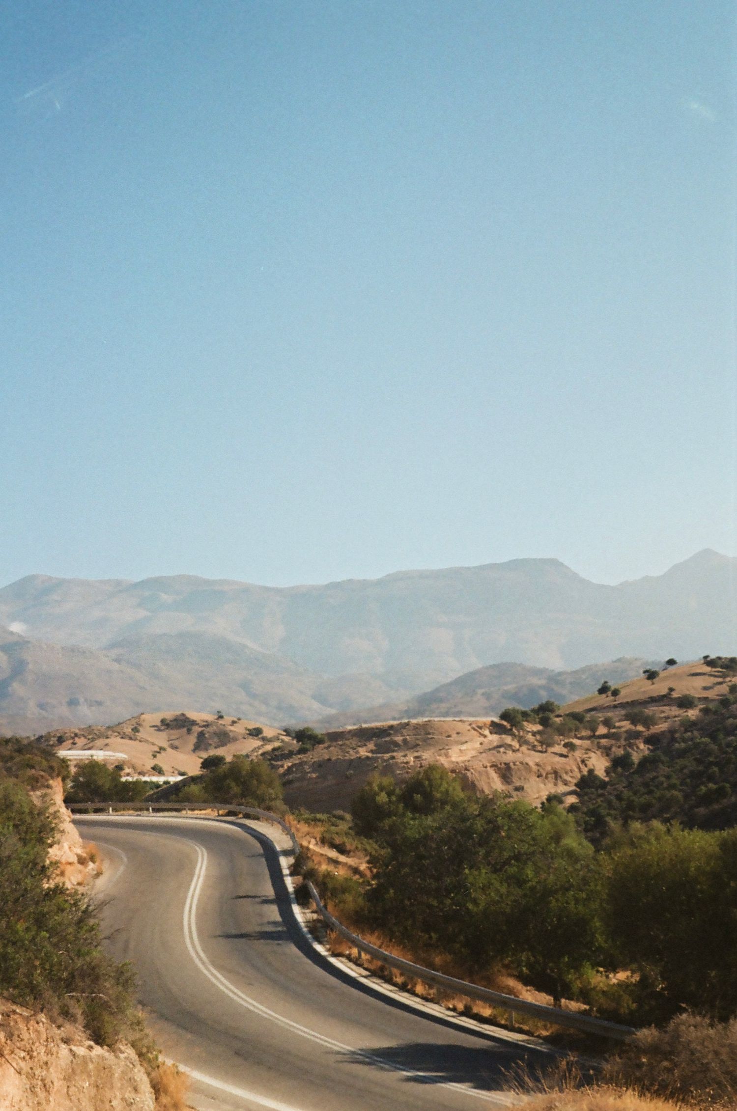 Winding mountain road curves through arid hills under clear blue sky in scenic landscape.