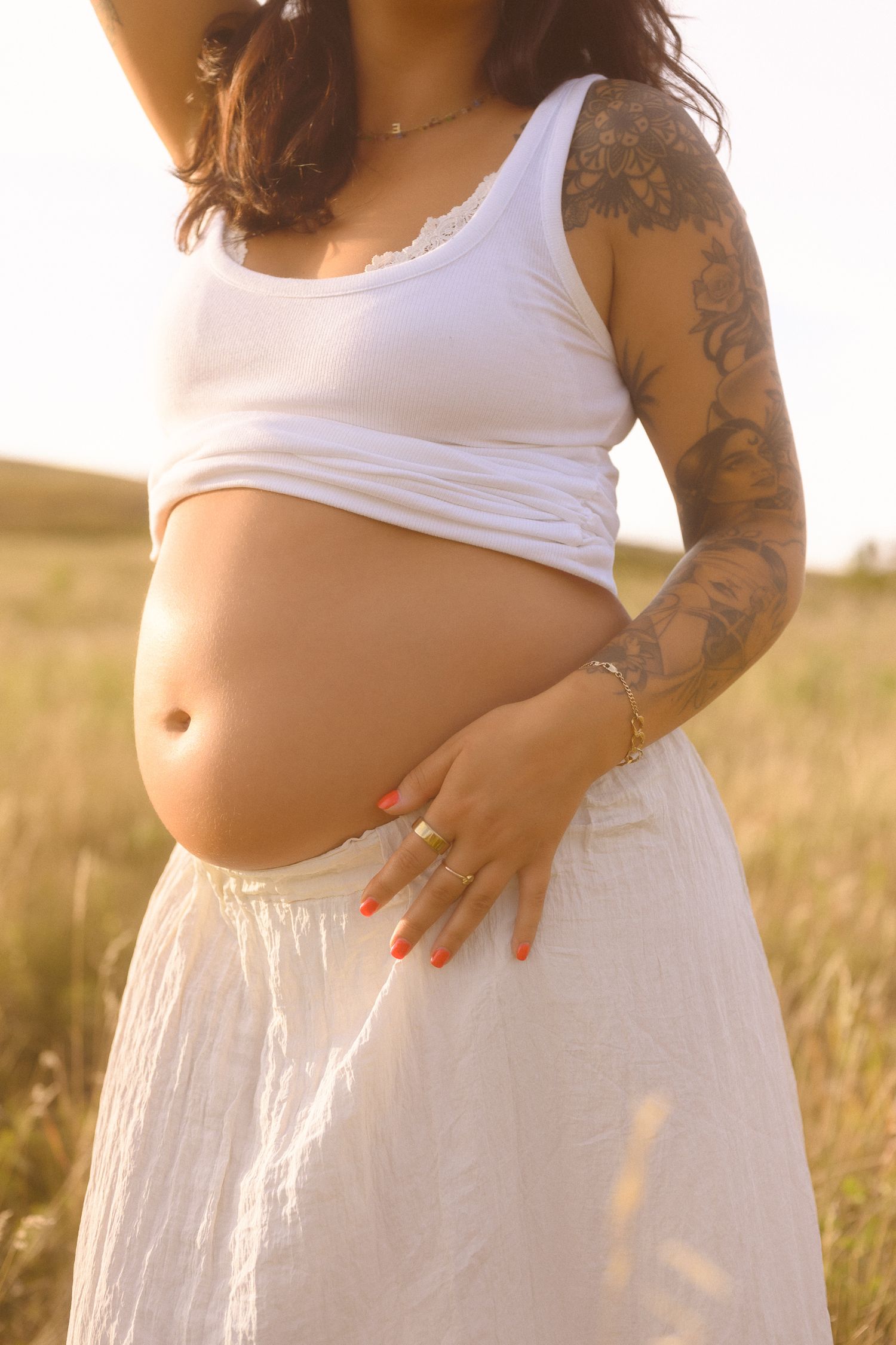 A maternity photoshoot in a sunlit field shows a person in white dress cradling their pregnant belly.