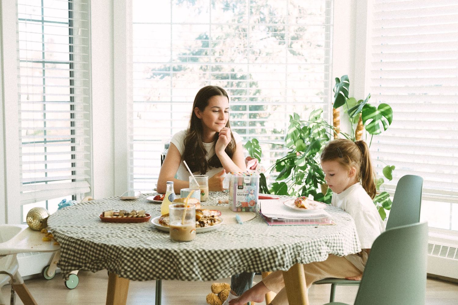Woman sitting at dining table with laptop and coffee cup in bright sunlit room with plants and modern decor.