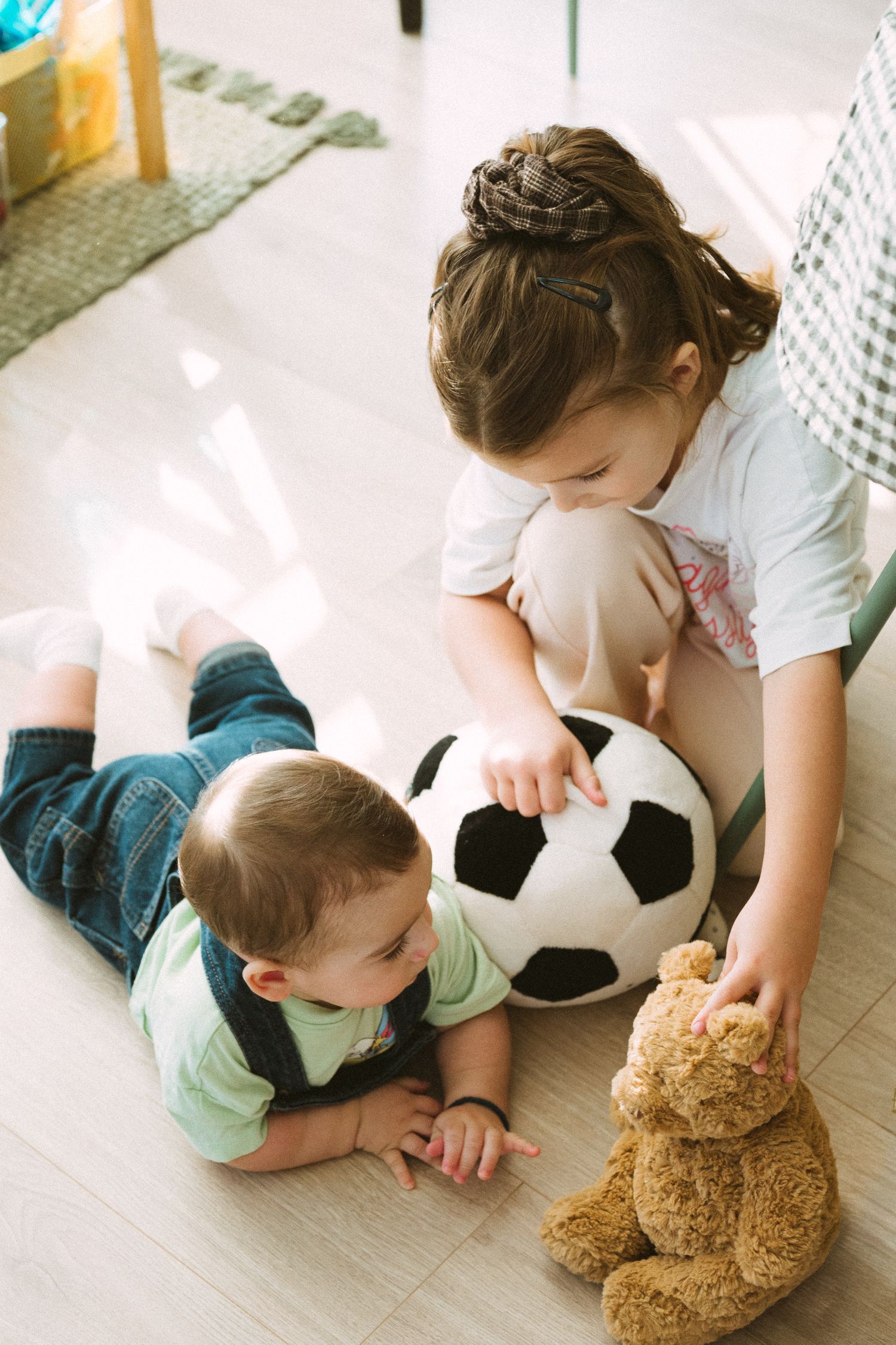 Children playing on the floor with a soccer ball and teddy bear in a bright sunlit room.