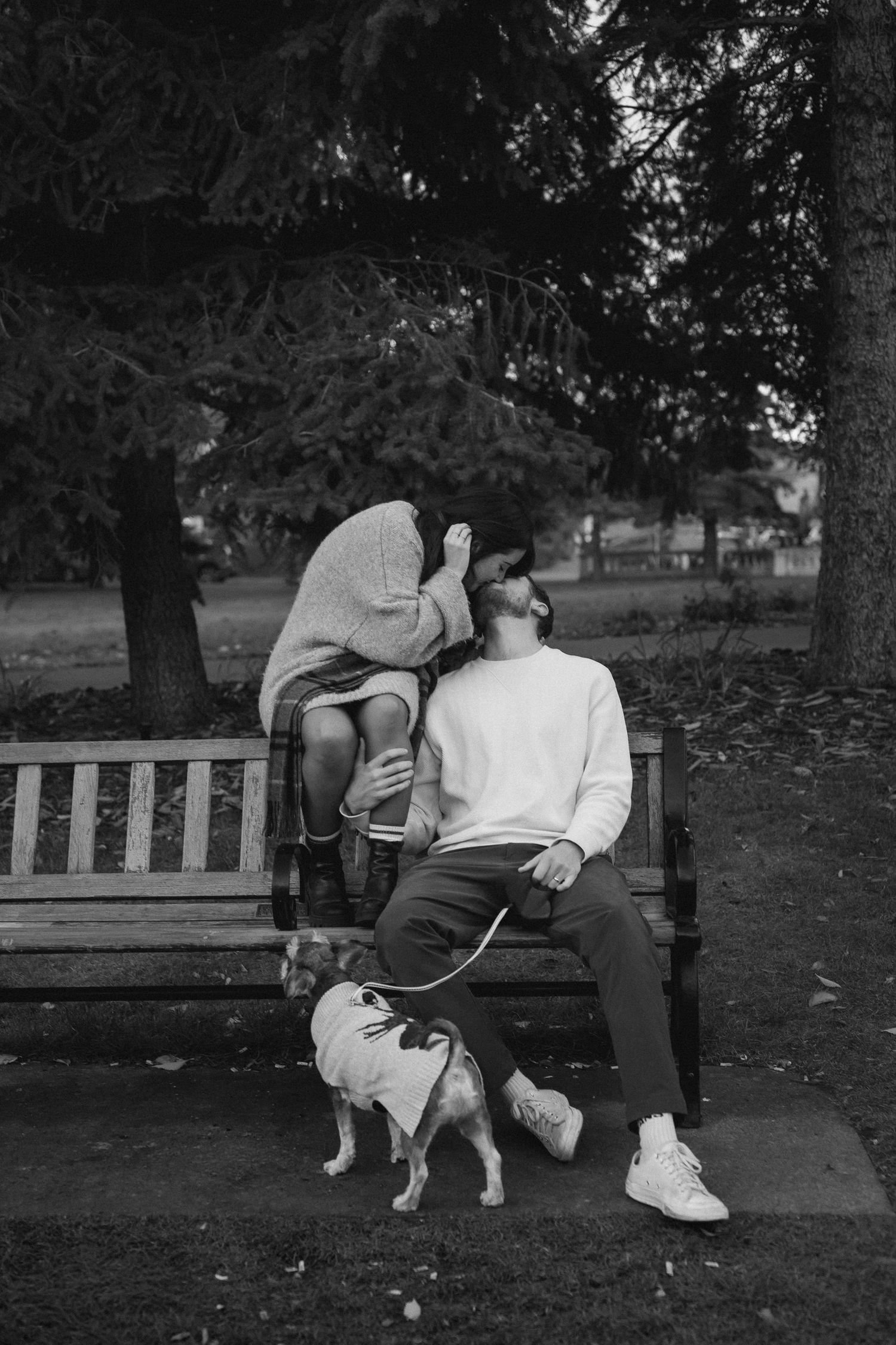 A couple sits together on a park bench in black and white, sharing an intimate moment in nature.