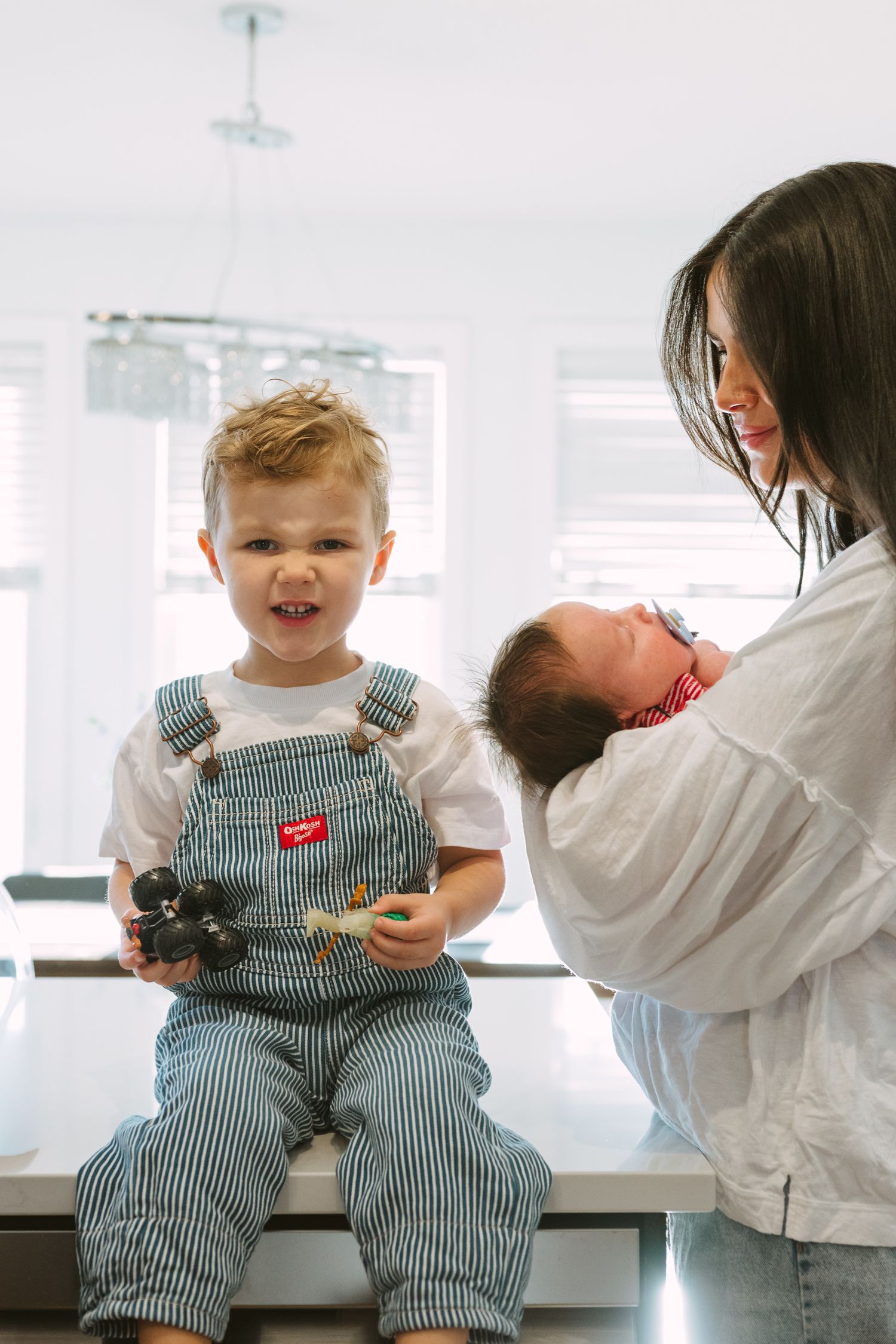 A child in striped overalls sits on a counter while someone holds a baby in a bright white kitchen setting.