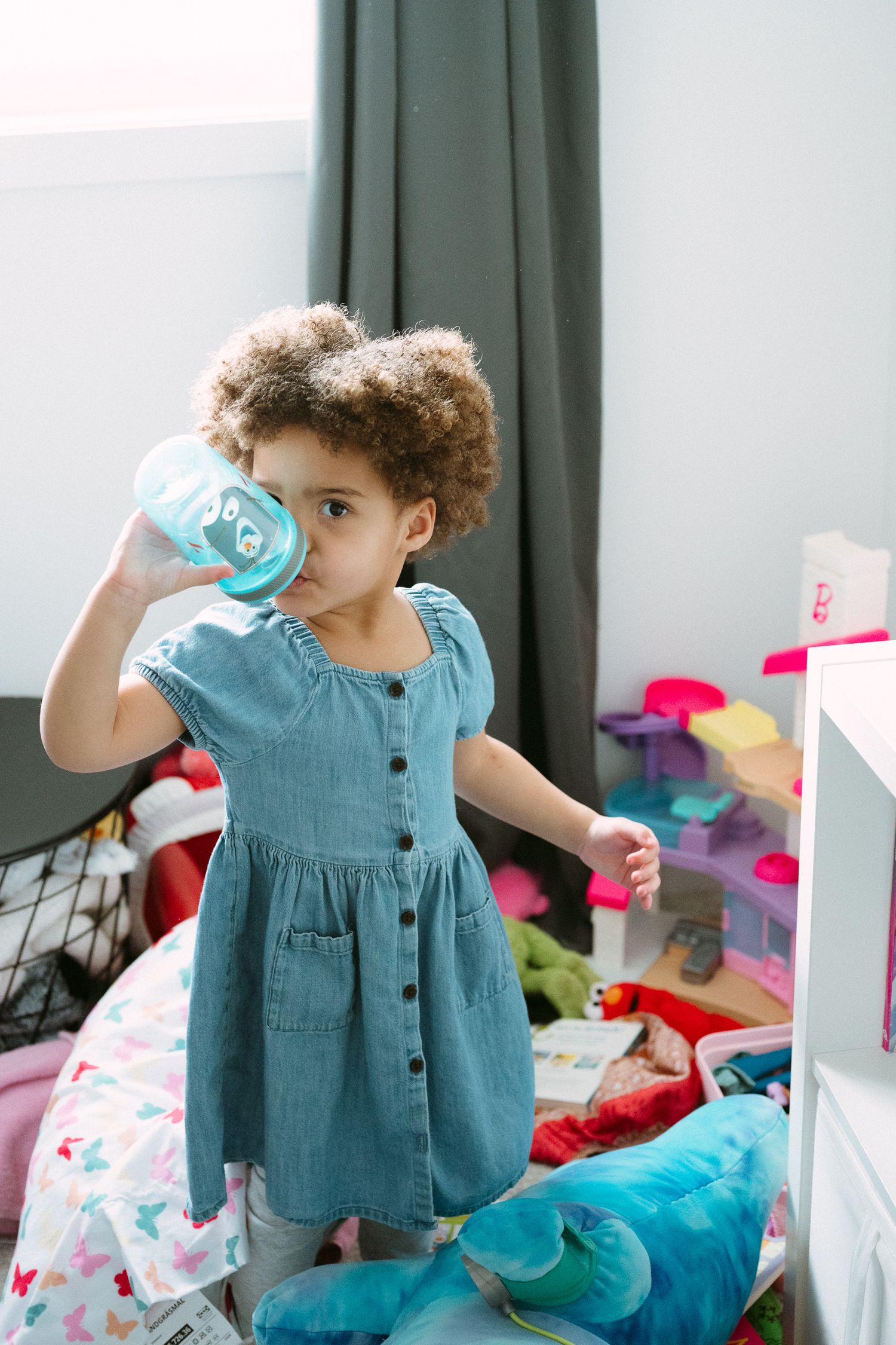 A curly-haired toddler girl in a denim dress drinks from a sippy cup in a colorful, toy-filled bedroom.