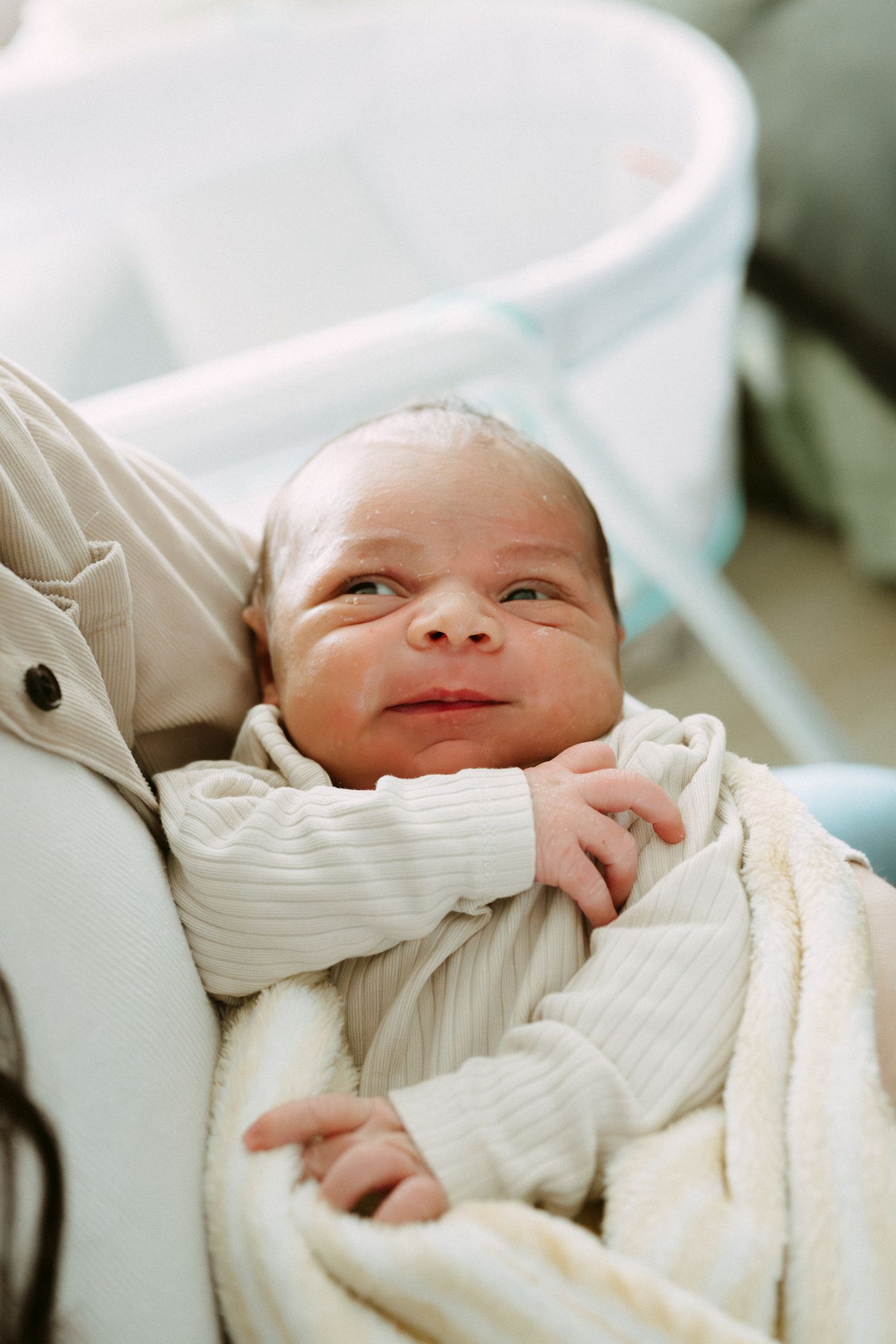 Newborn baby wrapped in cream blanket smiling while being held in hospital room.