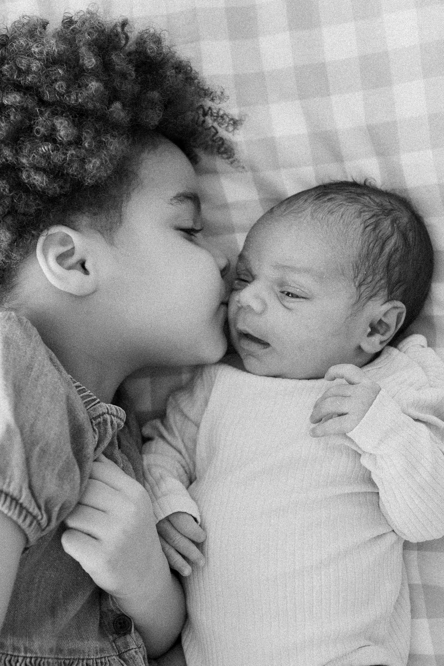 Black and white photo of a young child kissing a newborn baby lying on a gingham blanket.