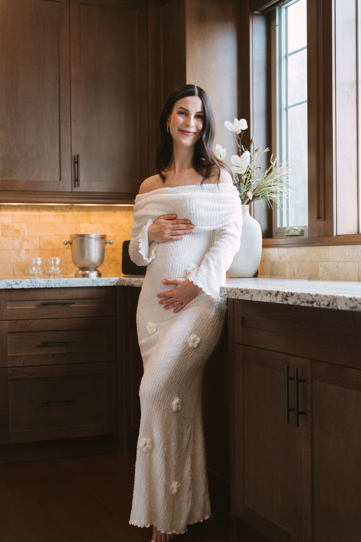 Smiling pregnant woman in white off-shoulder knit maternity dress posing in elegant kitchen with flowers.