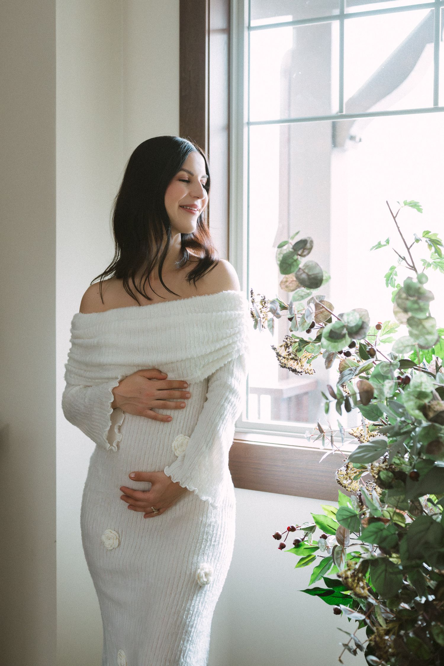 Smiling pregnant woman in white knit off-shoulder dress standing by a sunlit window with plants.