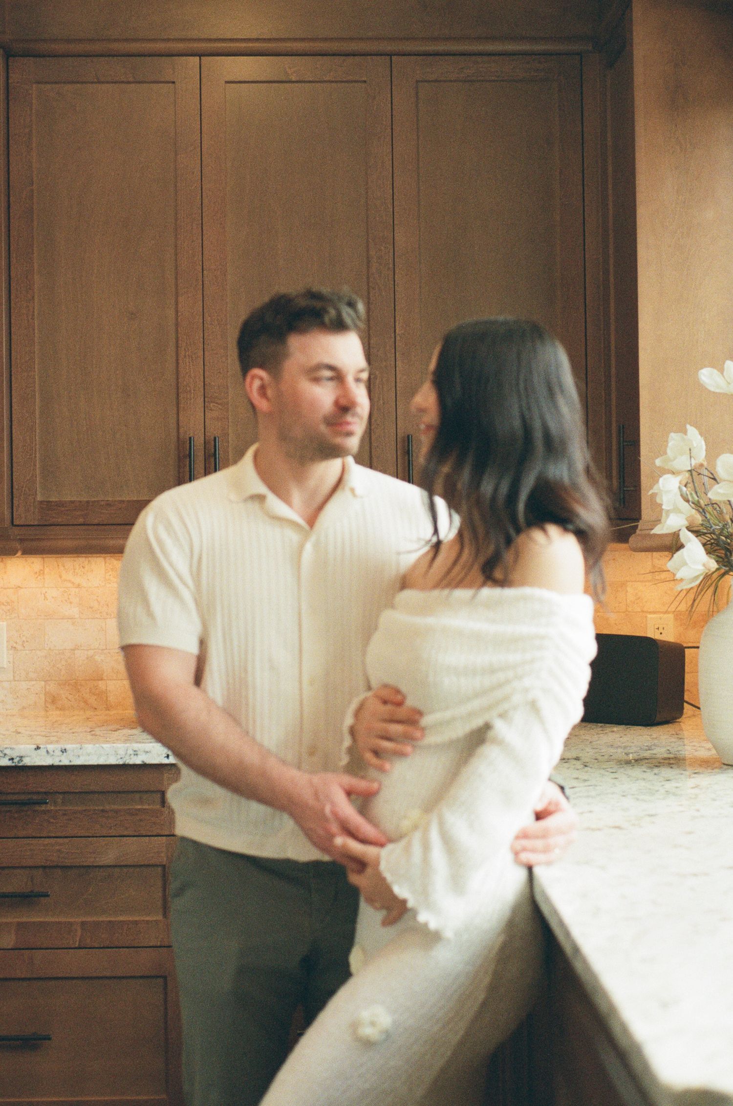 Couple sharing a tender moment in a warm kitchen, man holding pregnant woman in white off-shoulder dress.