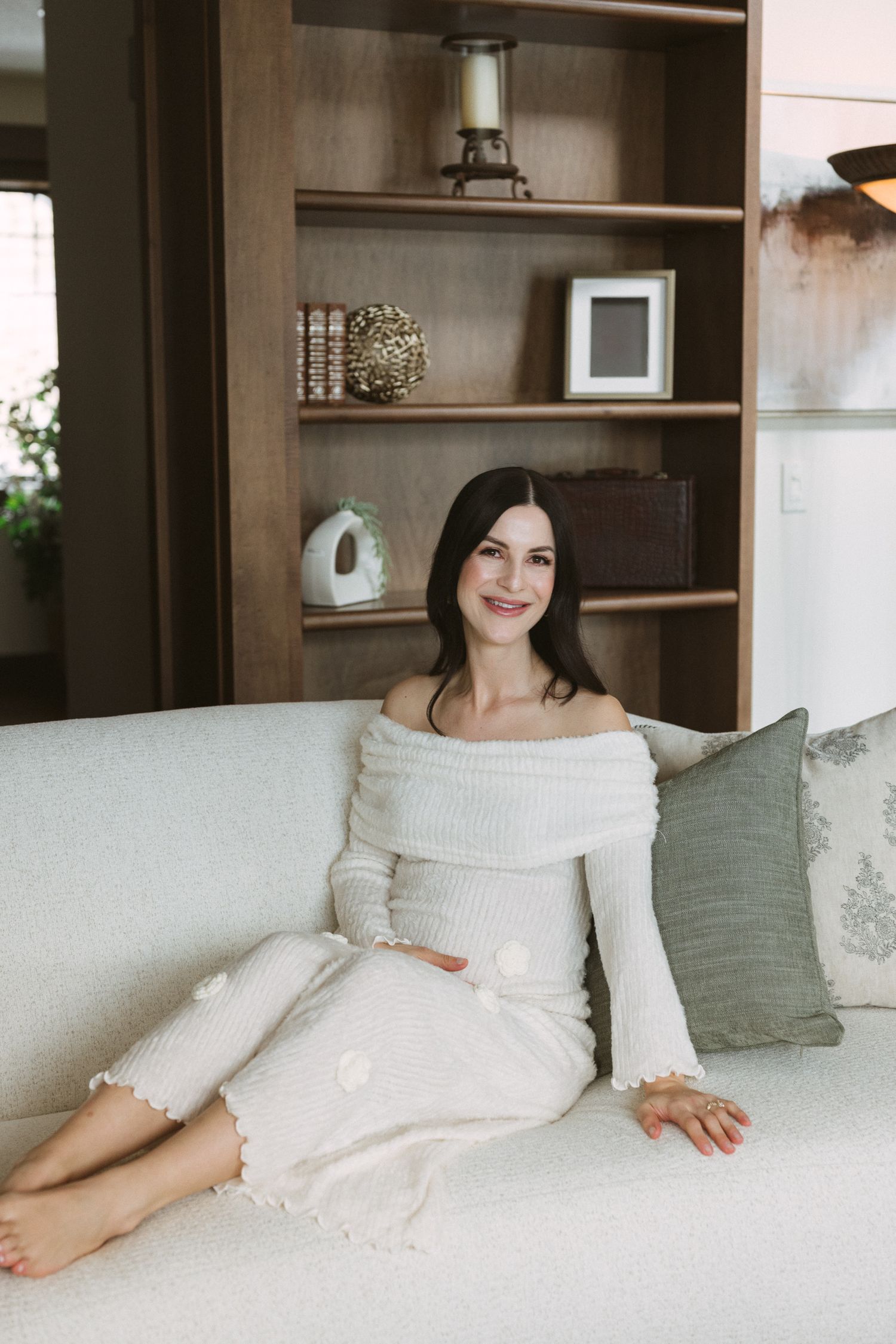 Smiling pregnant woman in white off-shoulder dress sitting relaxed on cream sofa in elegant living room.